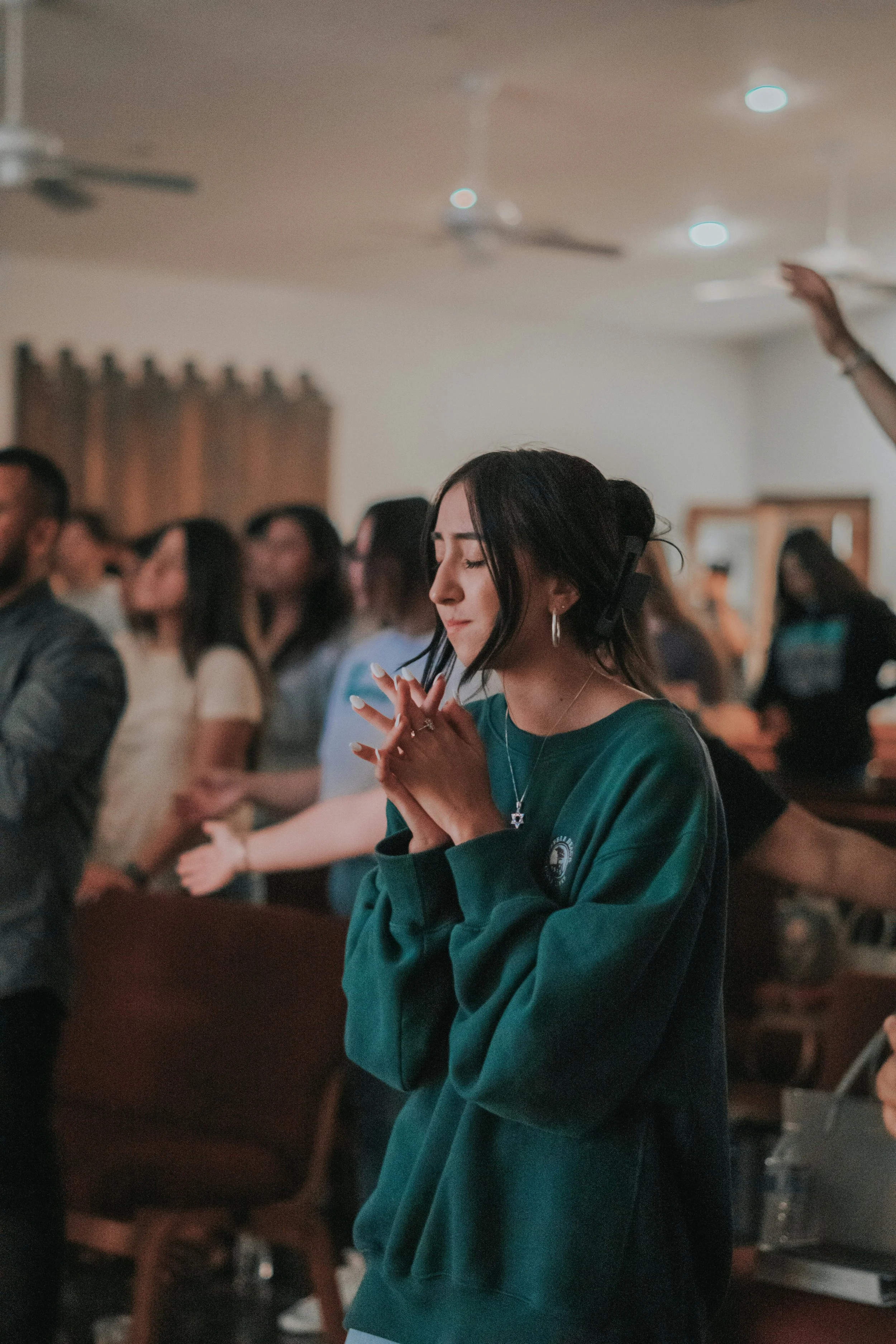 A young woman with dark hair, wearing a green sweatshirt, praying with her hands clasped and eyes closed in a church, surrounded by other people worshiping.