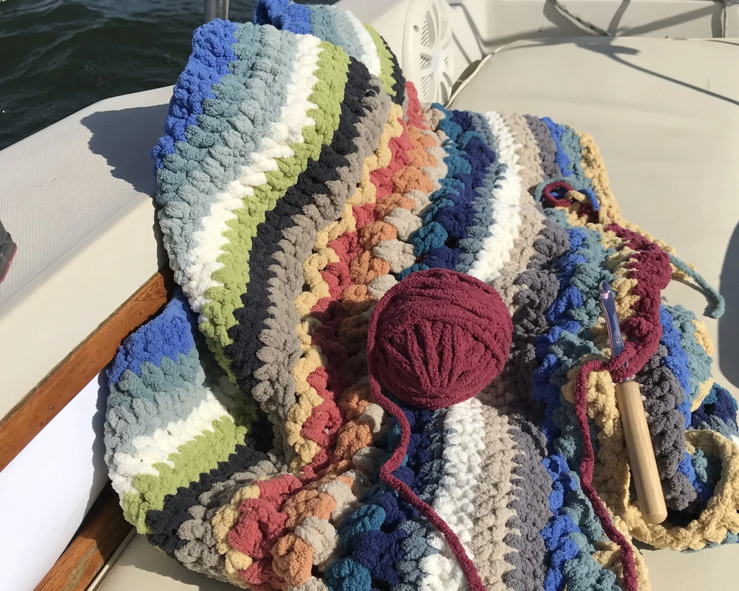 Colorful crocheted blanket on a boat with a ball of red yarn and a crochet hook.
