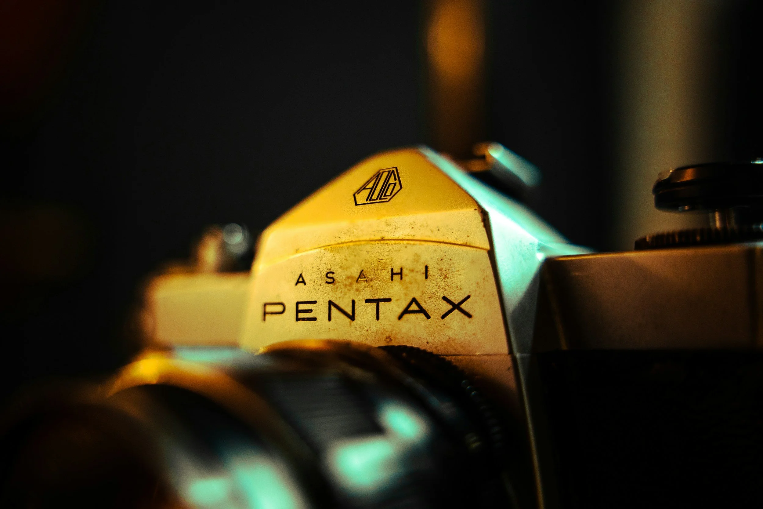 Close-up of a vintage camera with the brand name 'Pentax' and part of the logo visible, in a dark setting with a gold-colored section prominently displayed.