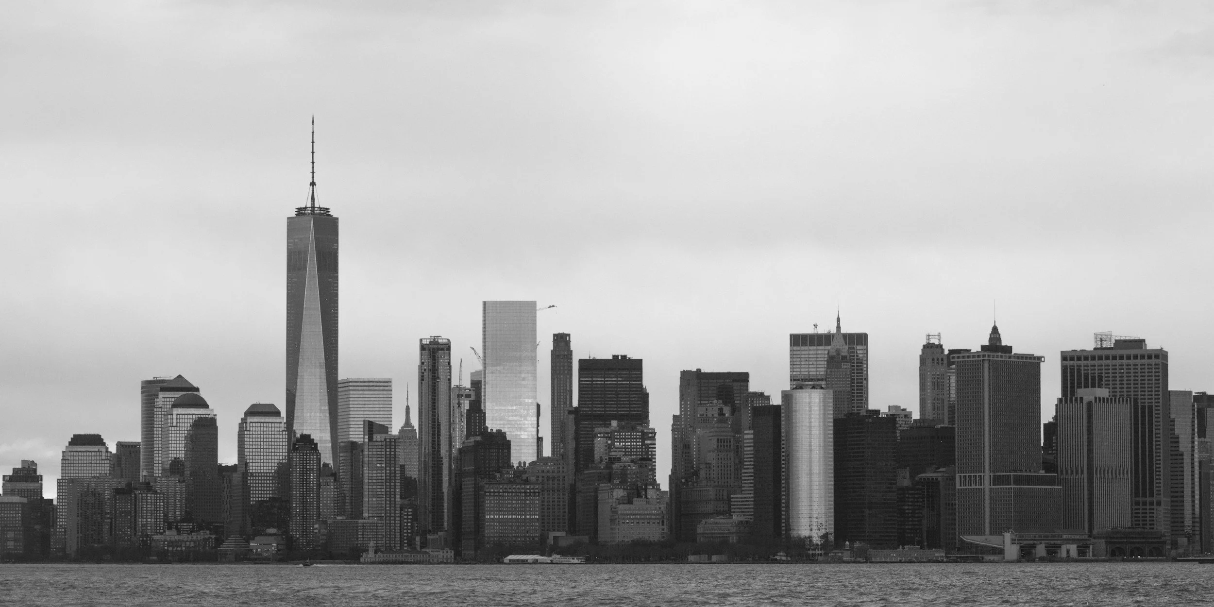 Black and white skyline of New York City with tall buildings, including One World Trade Center, viewed from across the water.