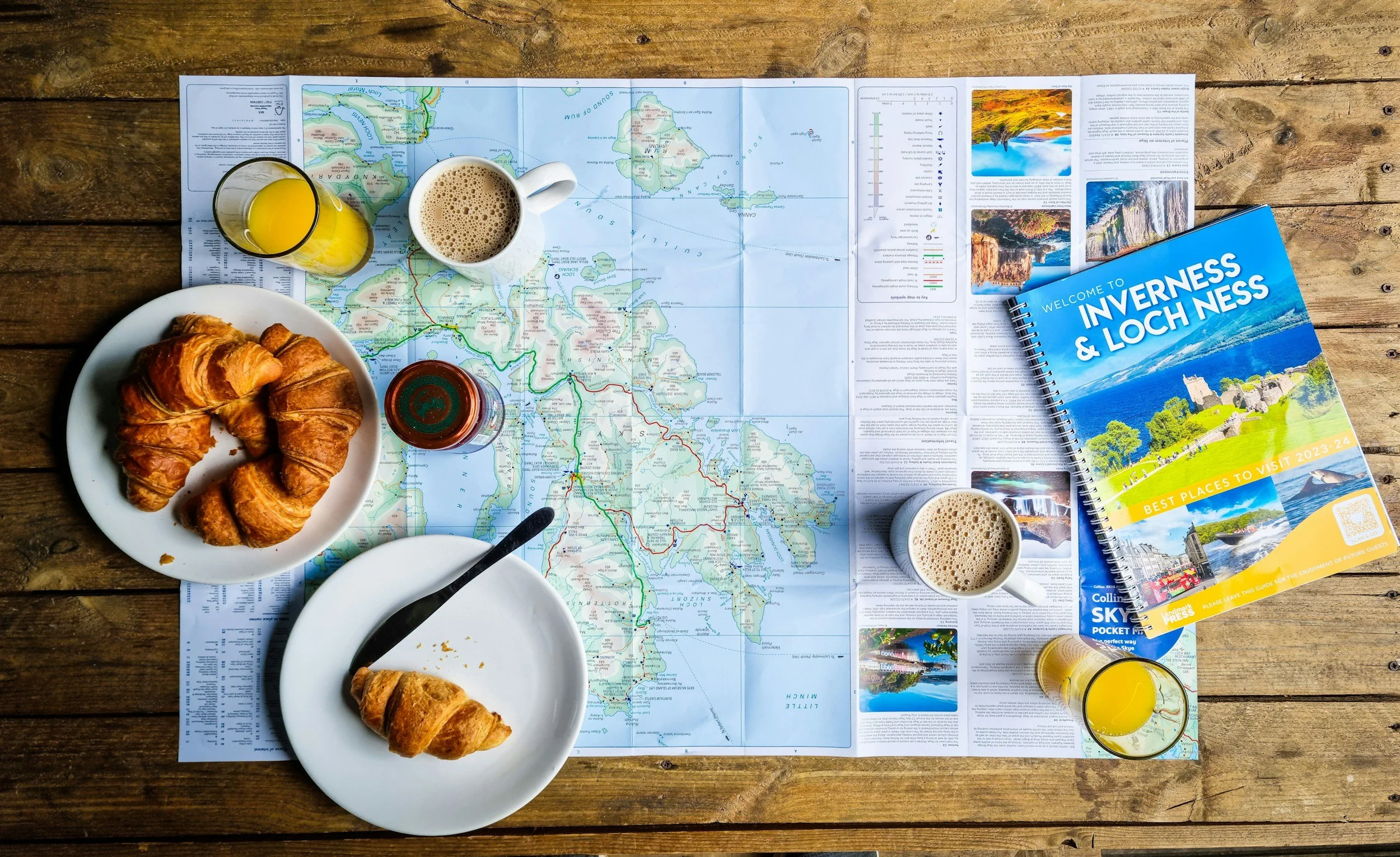 A map of the Isle of Skye in Scotland spread on a wooden table with two cups of coffee, a glass of orange juice, a white plate with three croissants, and a travel guidebook titled 'Welcome to Inverness & Loch Ness'.