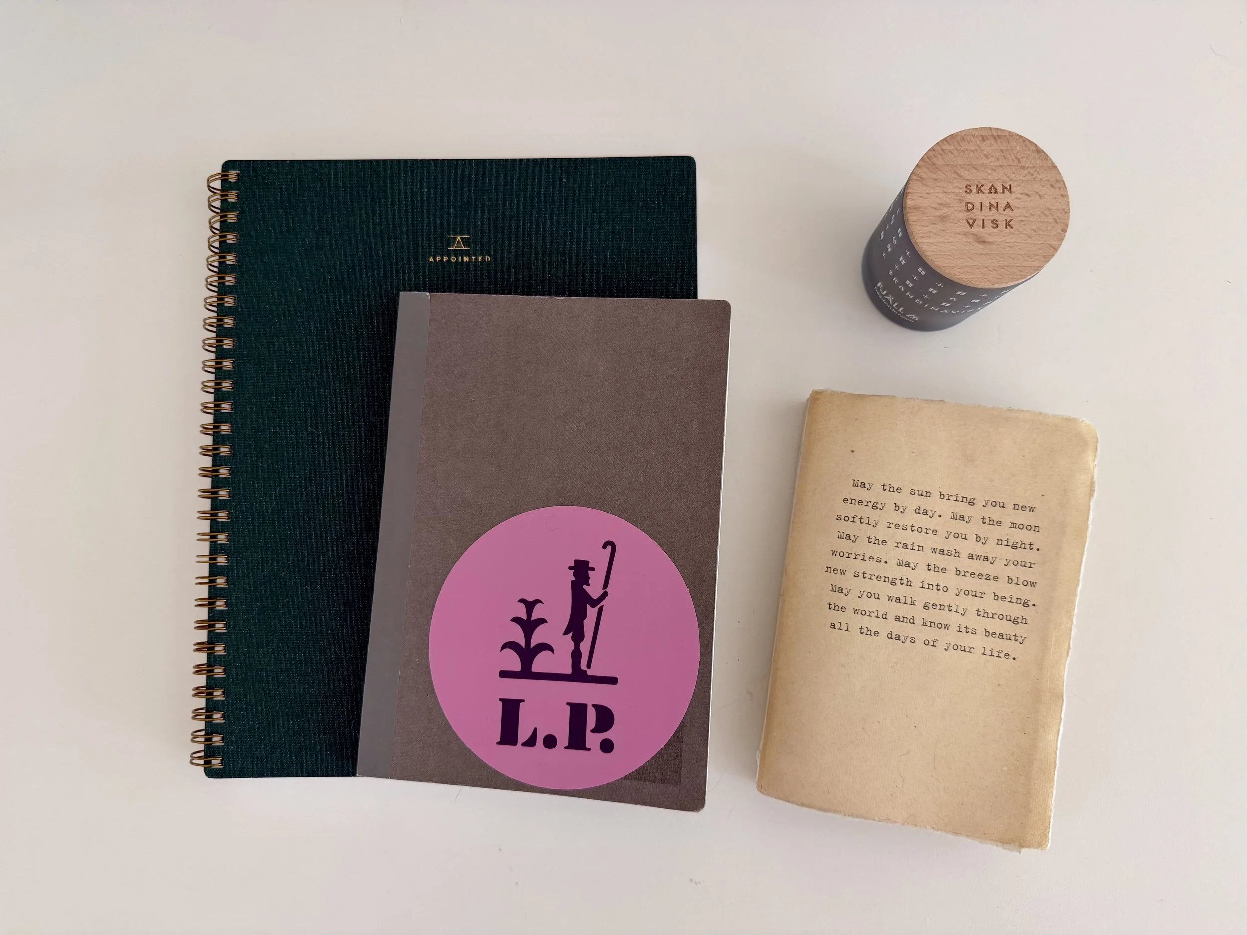 Top view of four notebooks and a printed paper on a white surface. The notebooks include a dark green spiral-bound, a gray one with a purple and pink sticker, and a black one with a wooden cap. The printed paper has a short inspirational message about well-being.