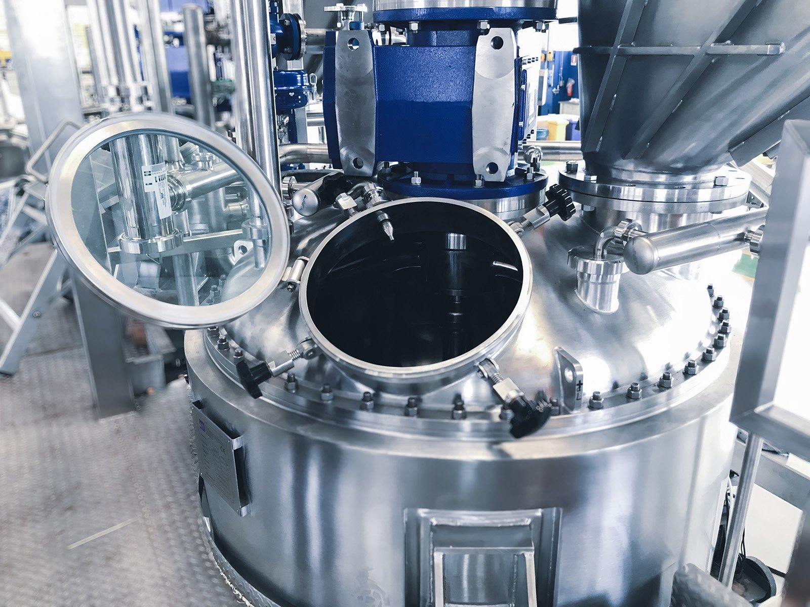 Industrial stainless steel equipment with circular opening, pipes, gauges, and a magnifying glass on the side, used in manufacturing or scientific processes.