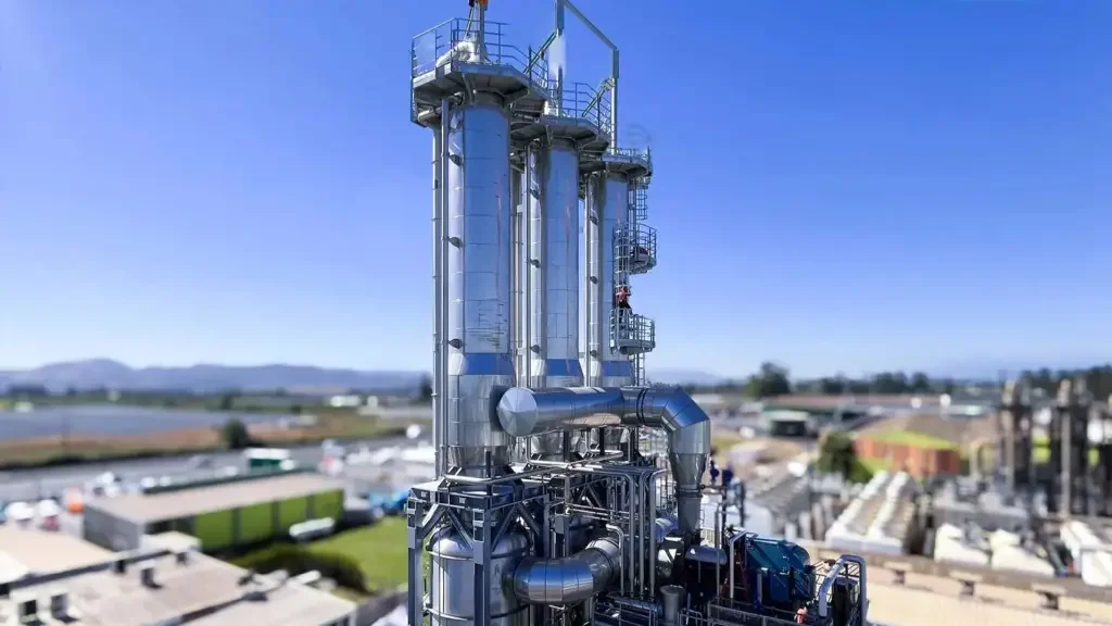 An industrial chemical processing tower with multiple interconnected pipes and ladders against a clear blue sky.