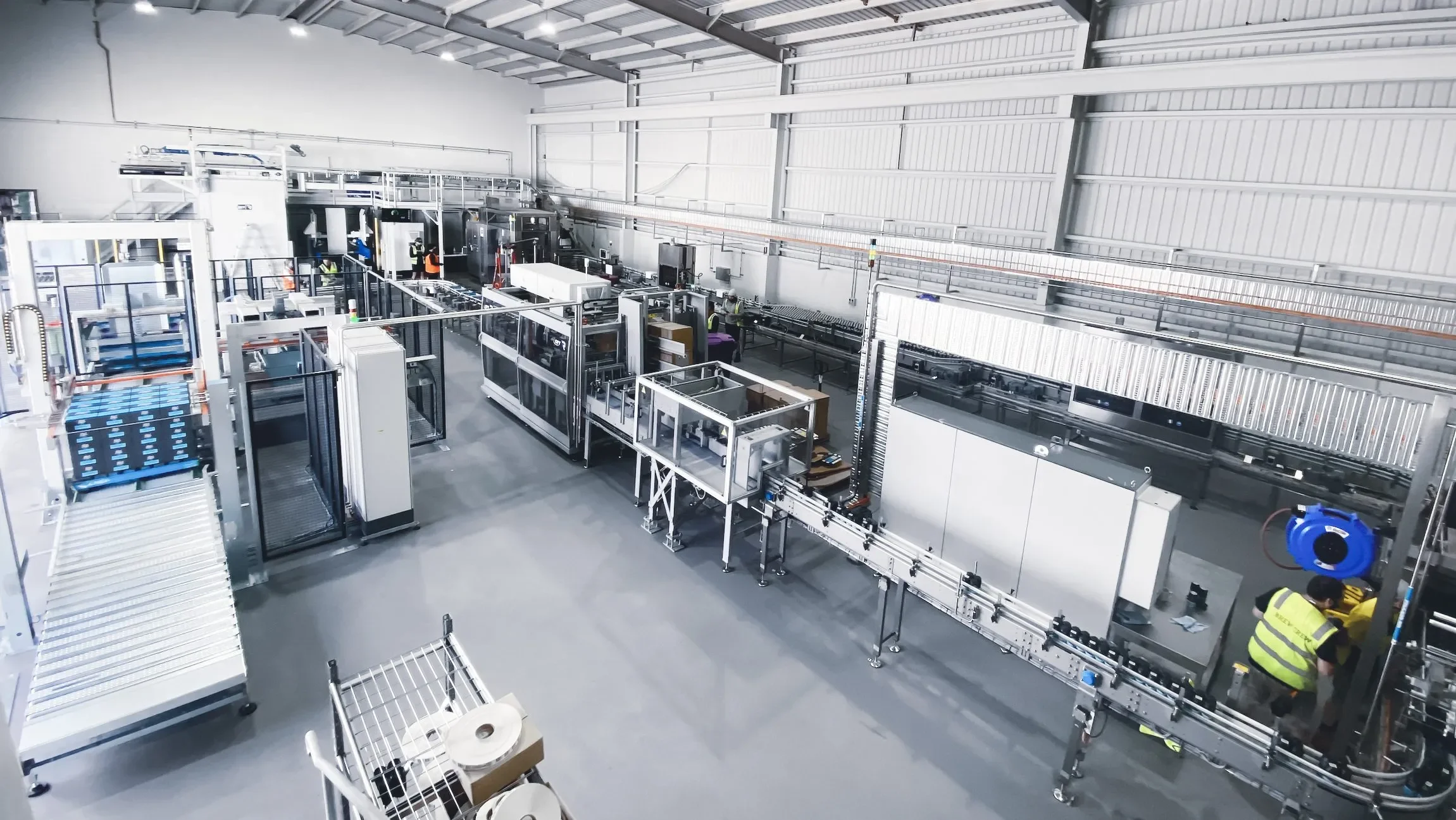 Interior view of an automated factory or warehouse with various conveyor belts, machines, and workers wearing safety vests.