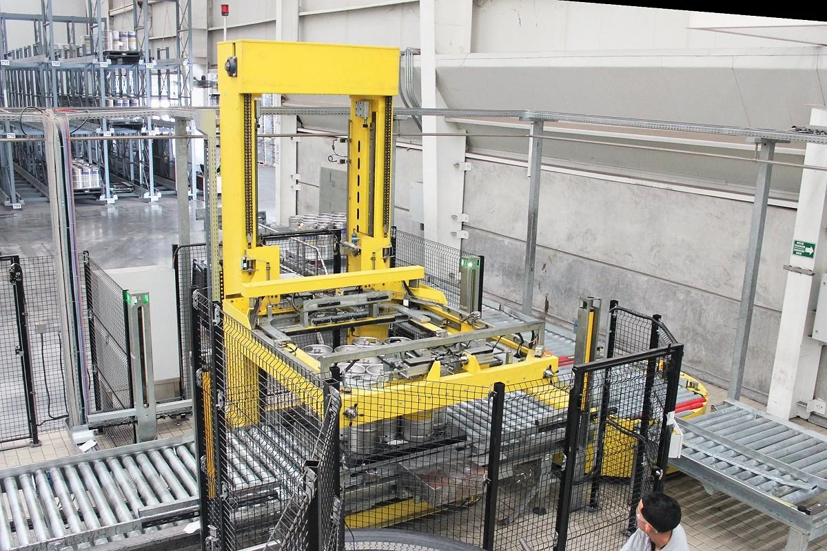 An industrial robotic machine with yellow and gray parts operating in a factory, surrounded by safety fences and conveyor belts.