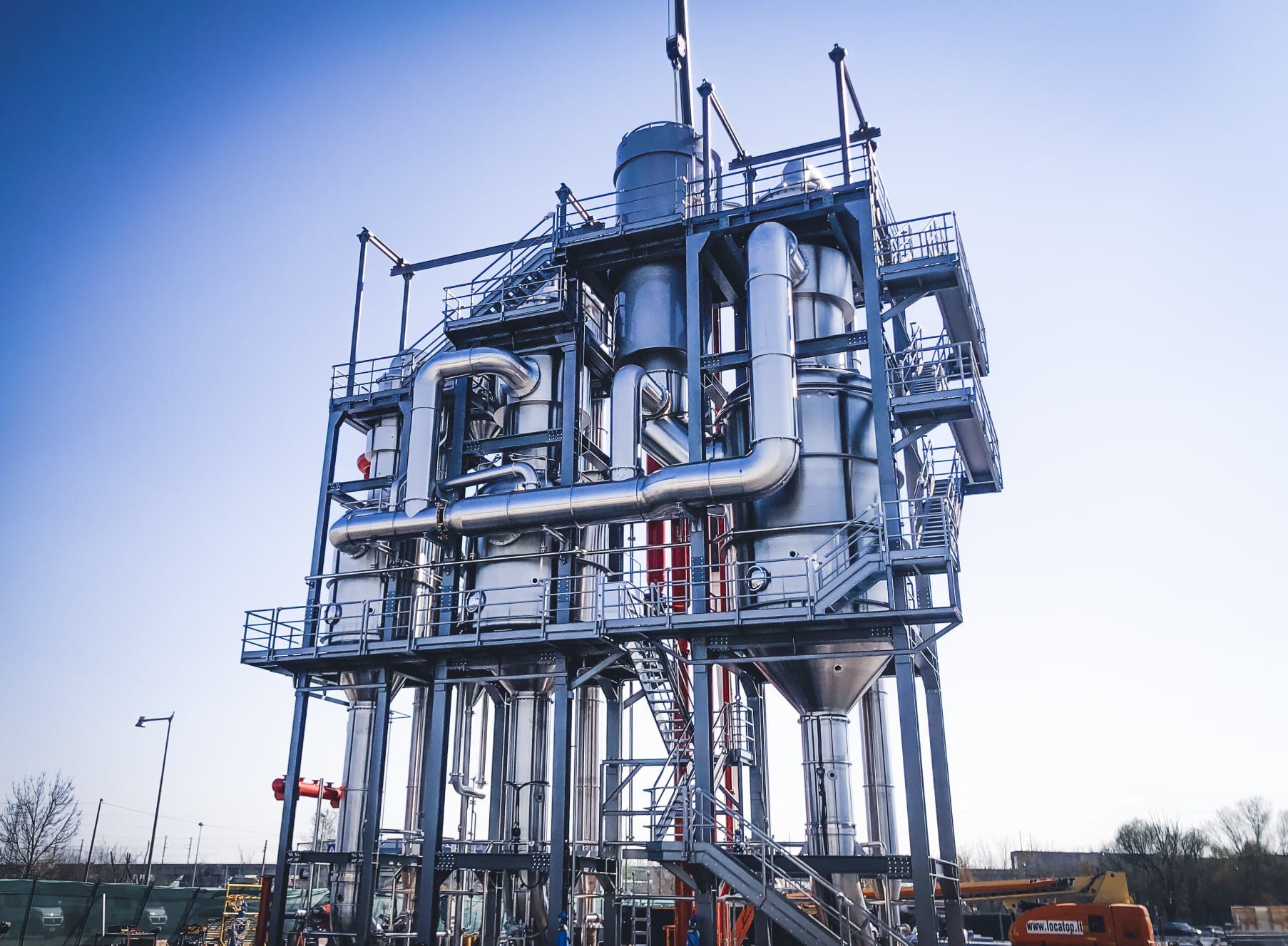 Industrial chemical processing plant with multiple metal pipes, tanks, and staircases under a clear sky.