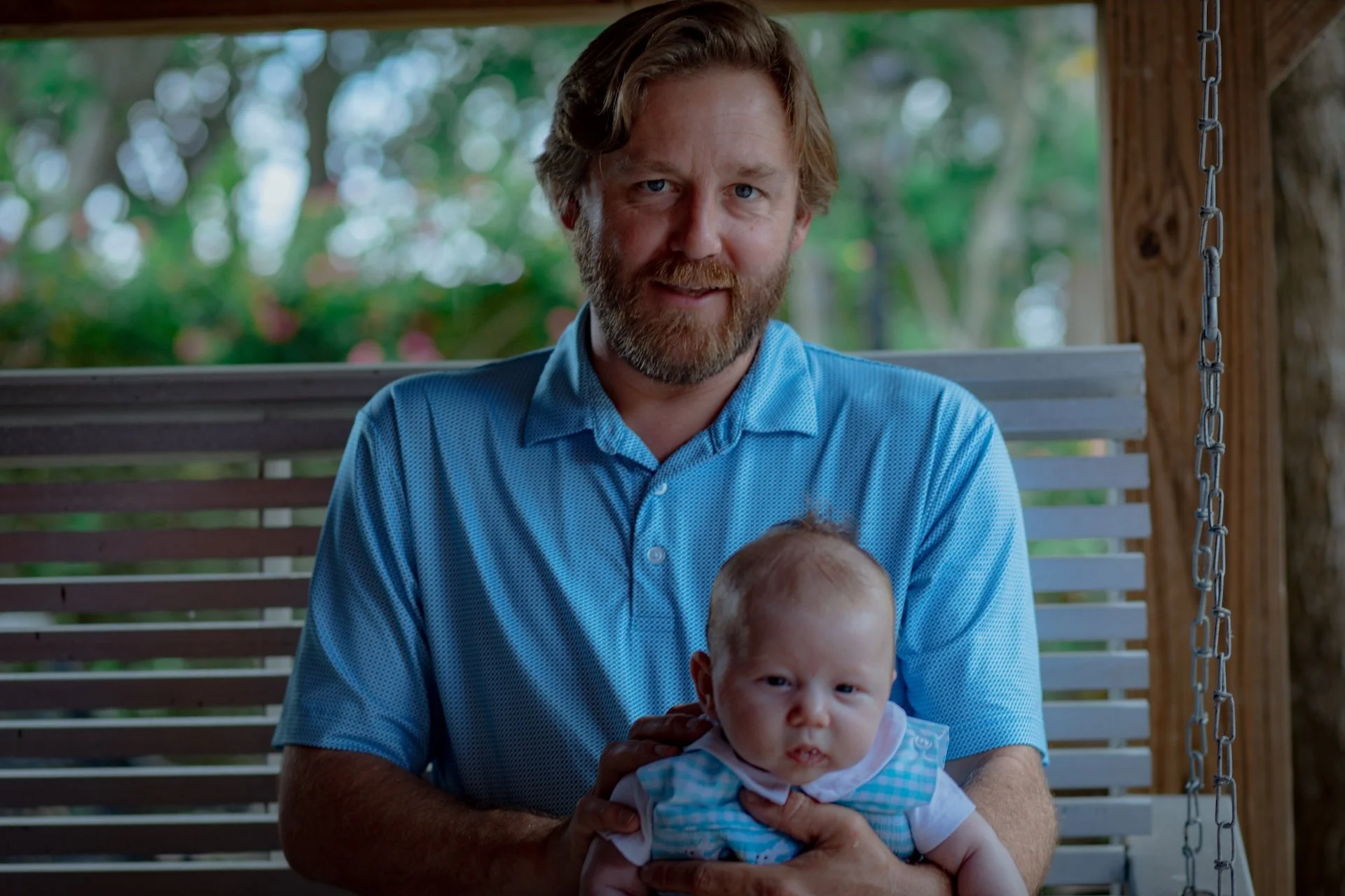 A man with a beard and light brown hair wearing a blue shirt sitting on a porch swing holding a young child with curly hair wearing a light blue shirt with white patterns. L'objectif Photography: New Orleans Wedding, Event, and Portrait Photographer
