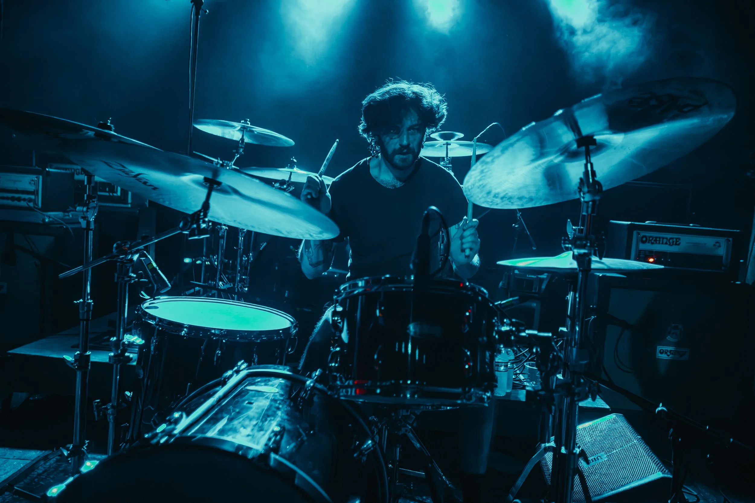 A drummer with curly hair playing drums on stage under blue lighting, with drums, cymbals, and electronic equipment visible.