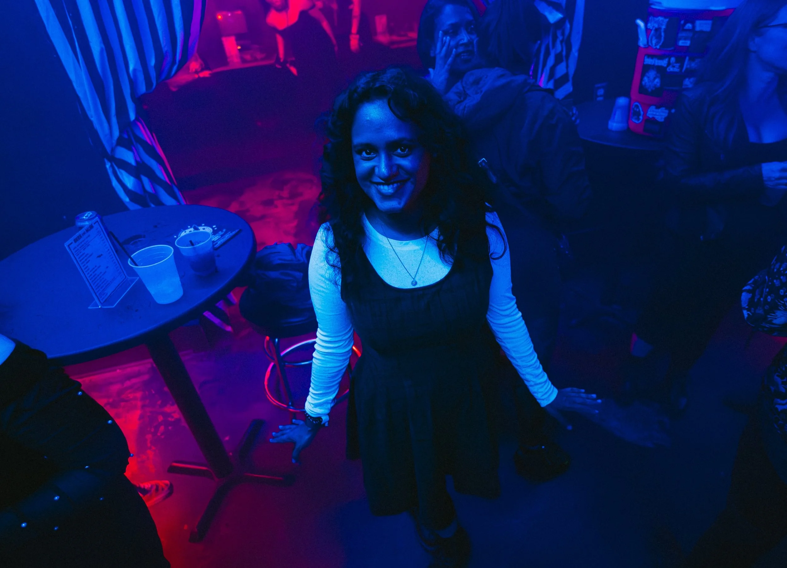 A woman with dark, curly hair smiling and looking up at the camera in a dimly lit bar or nightclub with colorful neon lighting. There is a table with drinks and utensils in front of her, and other people are visible in the background. L'objectif Phot