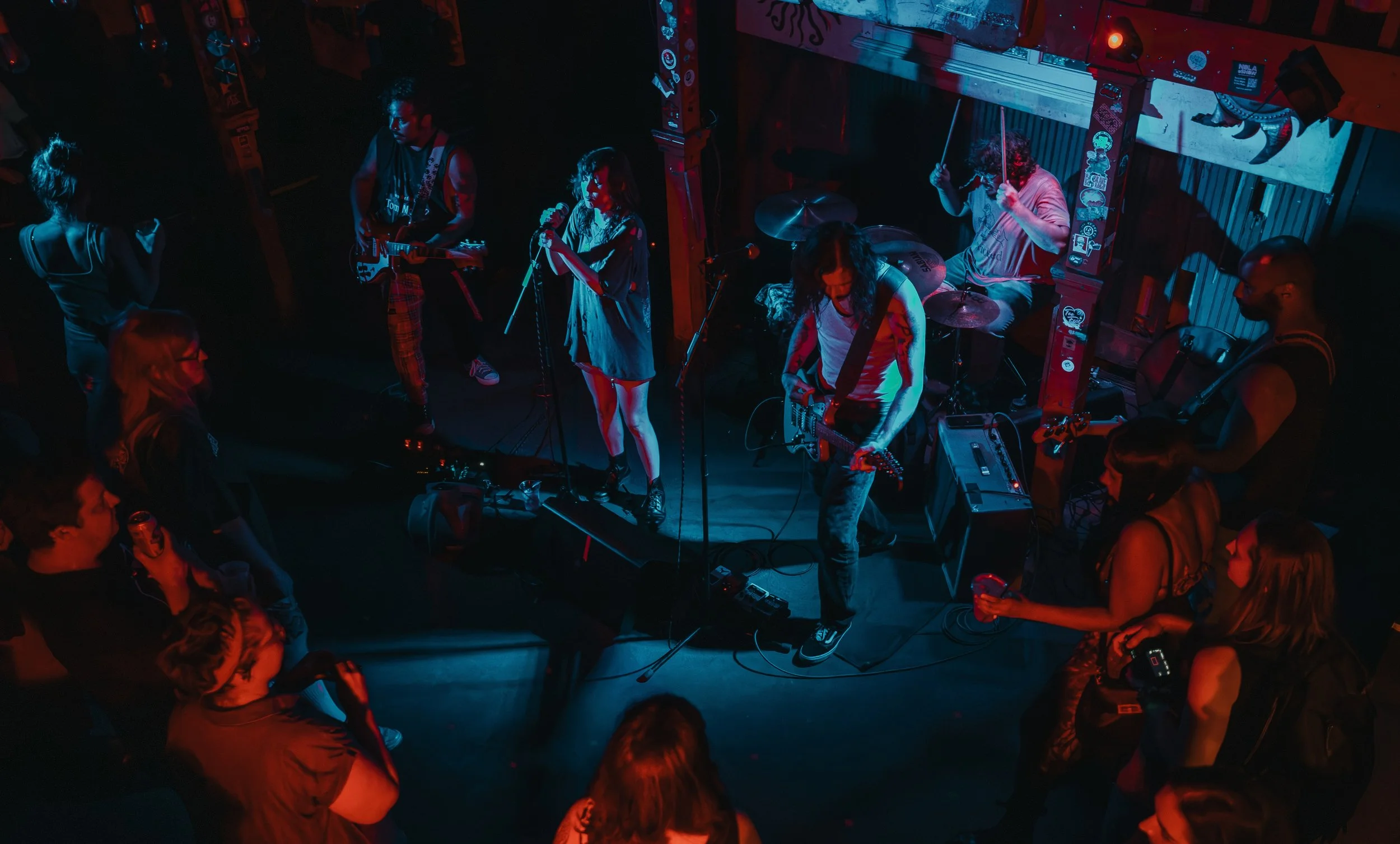 A band performing live on stage in a dimly lit venue with audience members watching, illuminated by colored stage lights. L'objectif Photography: New Orleans Wedding, Event, and Portrait Photographer