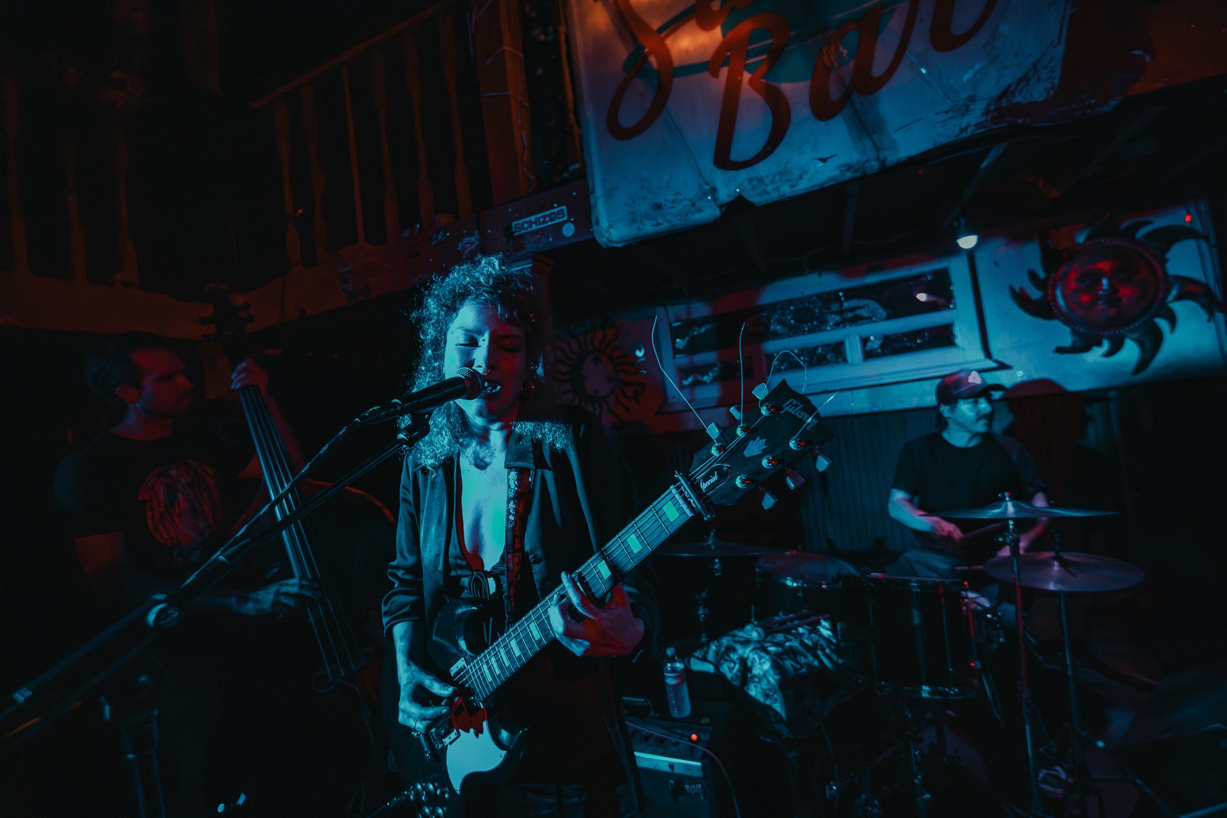 A band performing live on stage with a female guitarist singing into a microphone, illuminated by colorful stage lights, with a drummer playing in the background. L'objectif Photography: New Orleans Wedding, Event, and Portrait Photographer