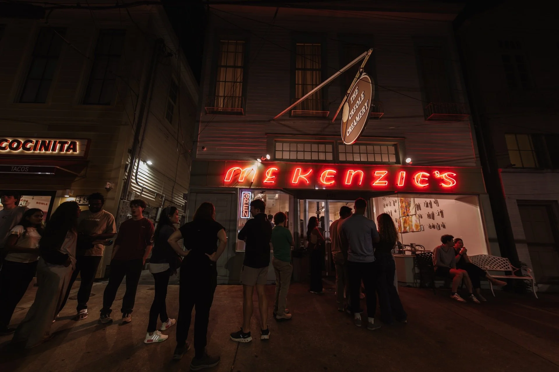 Nighttime scene outside McKenzie's with group of people waiting or socializing in front, neon sign of the restaurant illuminated, windows with warm light, and a marquee sign for The Cheesecake. L'objectif Photography: New Orleans Wedding, Event, and 