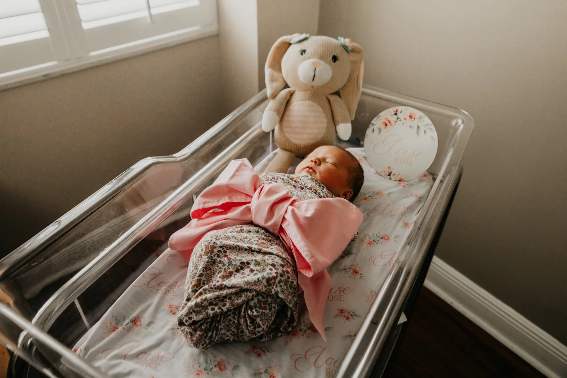 A newborn baby sleeping in a clear hospital bassinet, with a plush bunny stuffed animal next to them, and a decorative pillow with the words "Marise Rose" on it. L'objectif Photography: New Orleans Wedding, Event, and Portrait Photographer