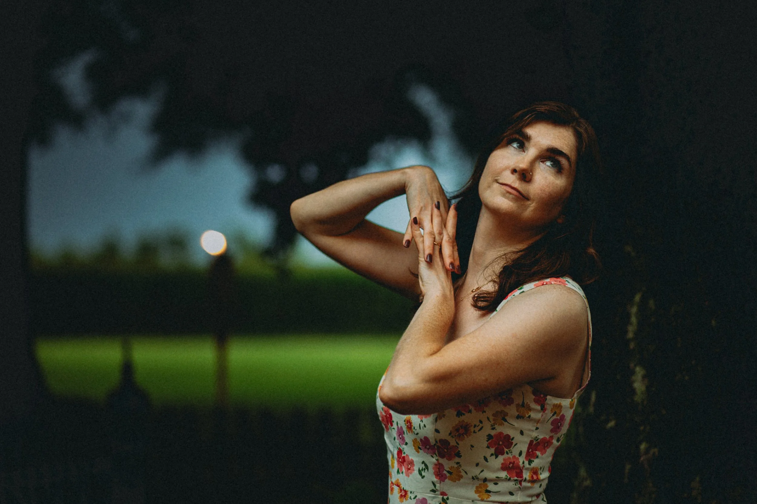A woman with red hair and a floral dress standing outdoors at dusk, with trees and a cloudy sky in the background. L'objectif Photography: New Orleans Wedding, Event, and Portrait Photographer
