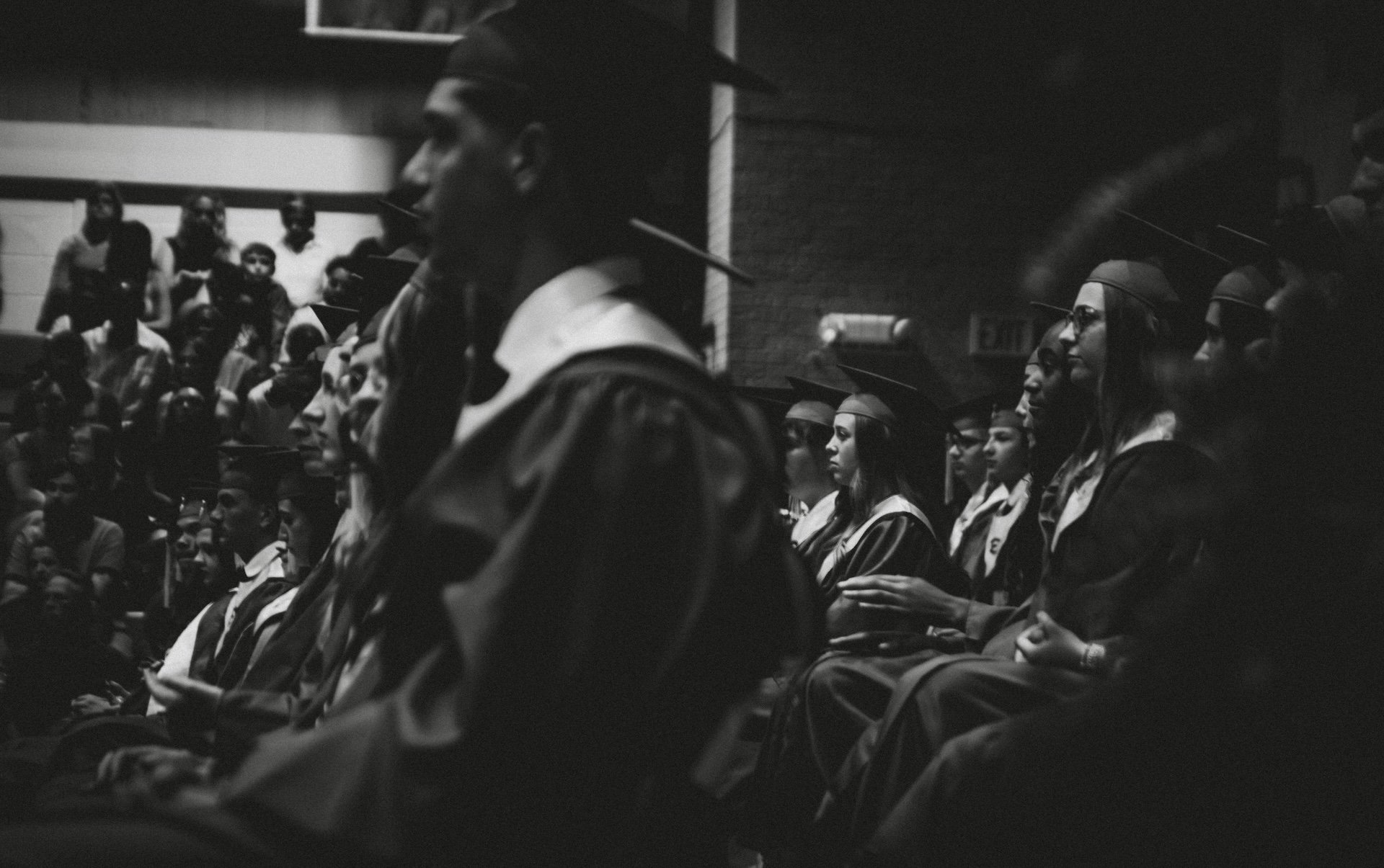 Black and white photo of people attending a graduation ceremony, wearing caps and gowns, sitting in rows, with some looking ahead and others reading or using devices. L'objectif Photography: New Orleans Wedding, Event, and Portrait Photographer