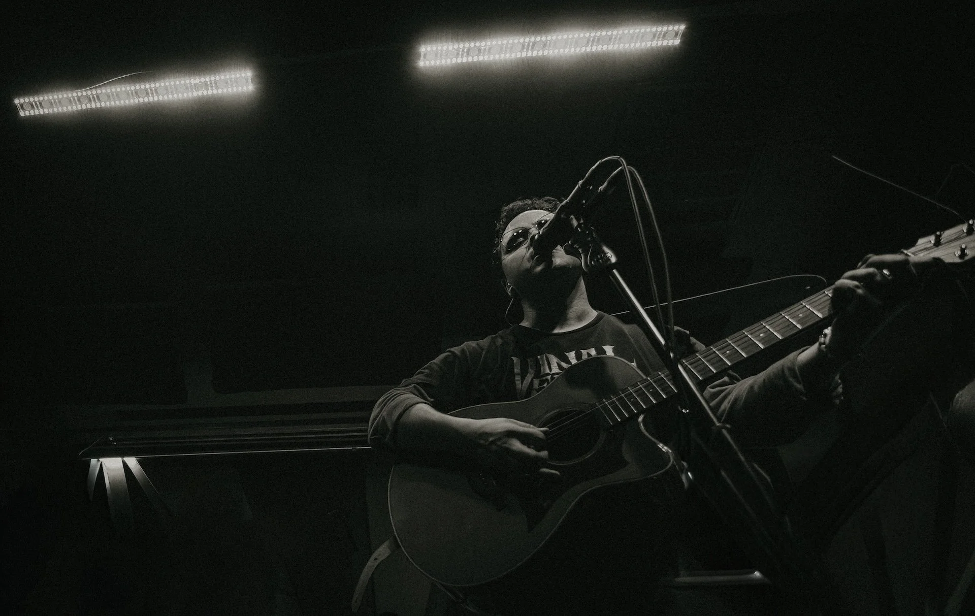 A person wearing sunglasses and a T-shirt is playing an acoustic guitar and singing into a microphone in a dimly lit room with minimal lighting. L'objectif Photography: New Orleans Wedding, Event, and Portrait Photographer