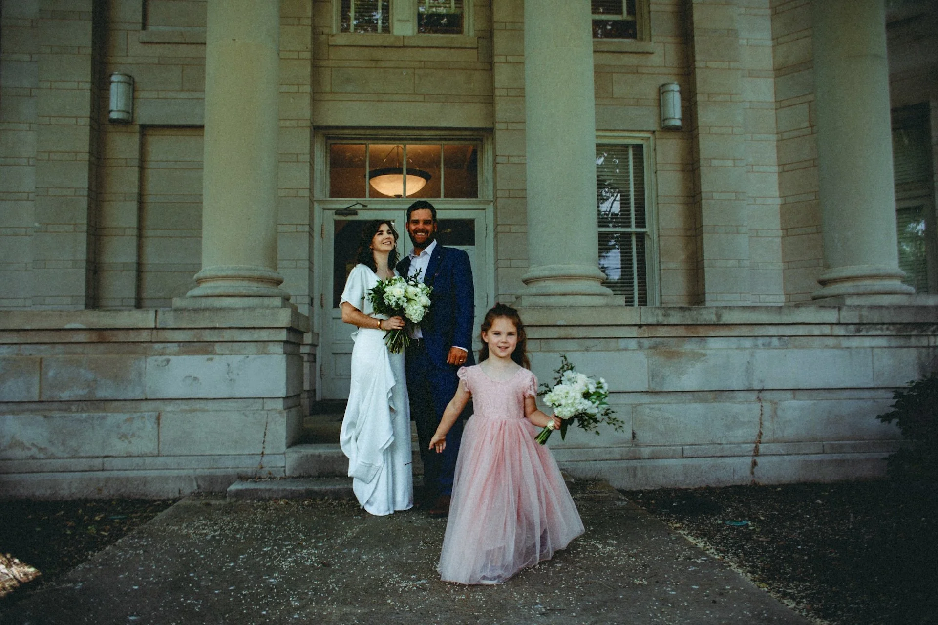 A happy newlywed couple and a young girl holding bouquets and standing in front of a large stone building with columns, during daylight. L'objectif Photography: New Orleans Wedding, Event, and Portrait Photographer