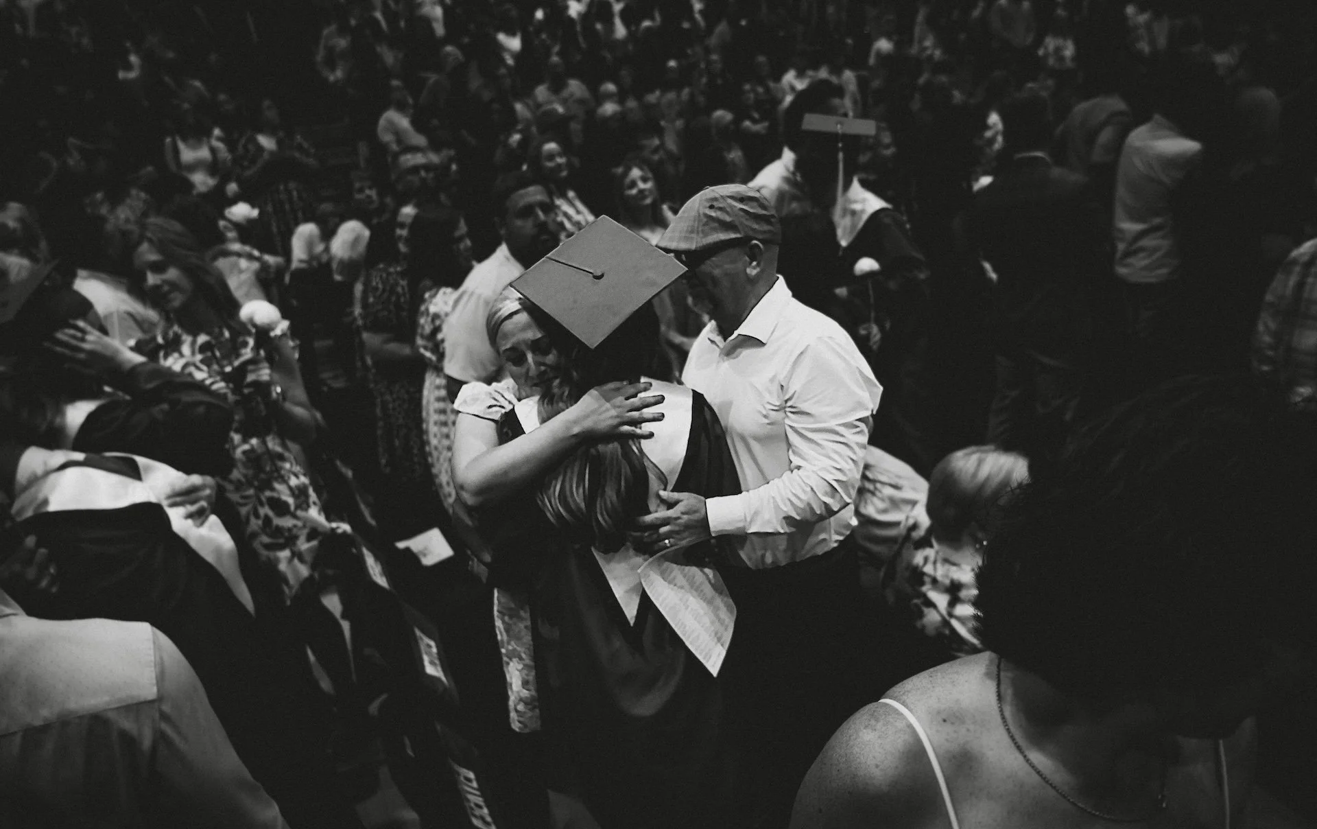 A black and white photo of a crowded graduation ceremony, showing a graduate hugging an older man, with many other attendees in the background. L'objectif Photography: New Orleans Wedding, Event, and Portrait Photographer