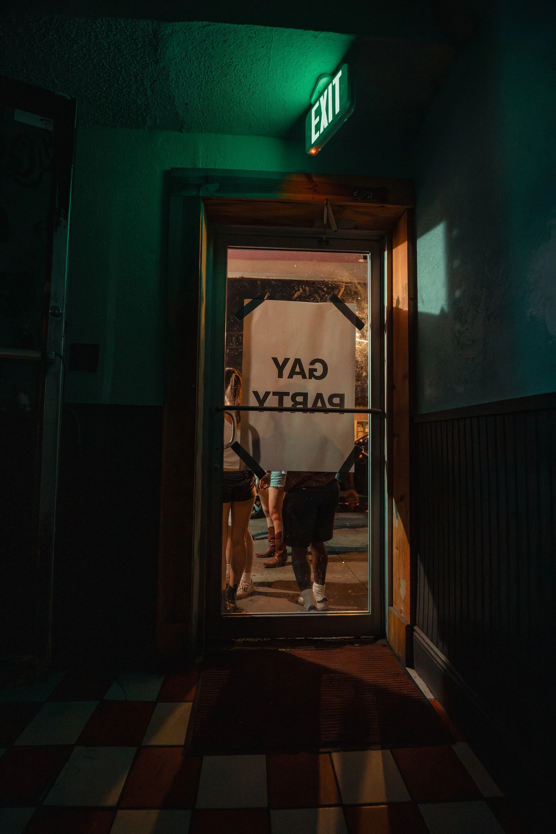 People standing near the entrance of a bar or club, with a sign on the door that reads 'Bay Party.' The doorway is open, and the interior is dimly lit.