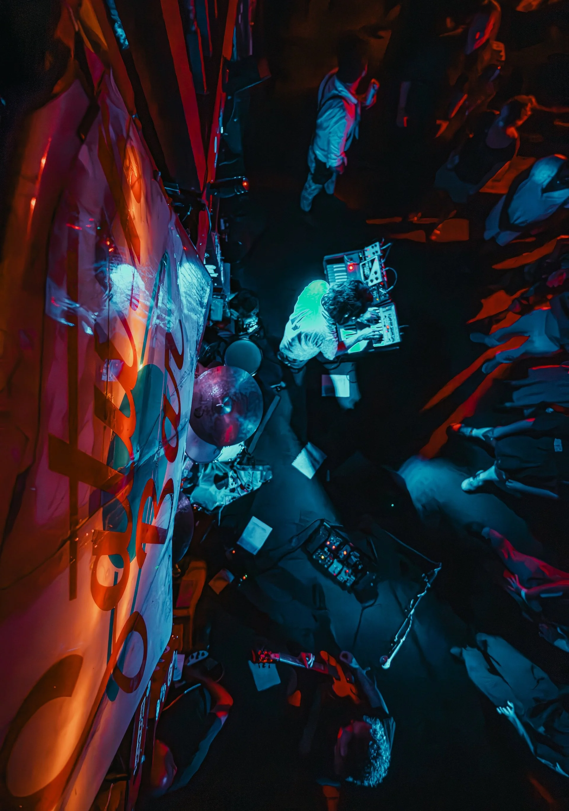 A DJ performing in a dark room with neon lighting, surrounded by a crowd of people, some standing and some sitting, with music equipment and a drum set visible. This could be a promo shot available for a band playing in New Orleans.
