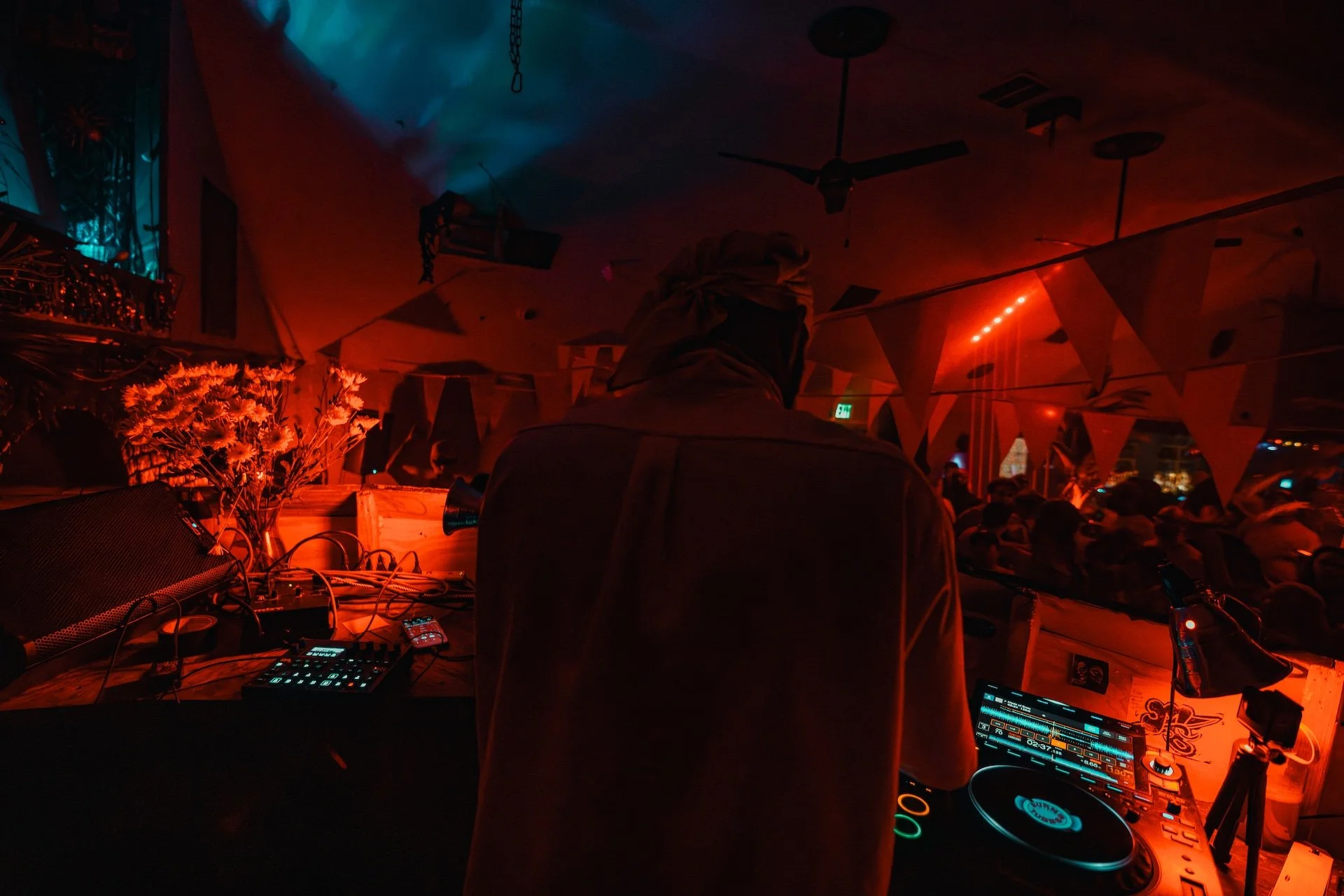 A DJ performing at a nightclub with red and blue lighting, some bunting decorations hanging on the ceiling, and a crowd of people in the background. L'objectif Photography: New Orleans Wedding, Event, and Portrait Photographer