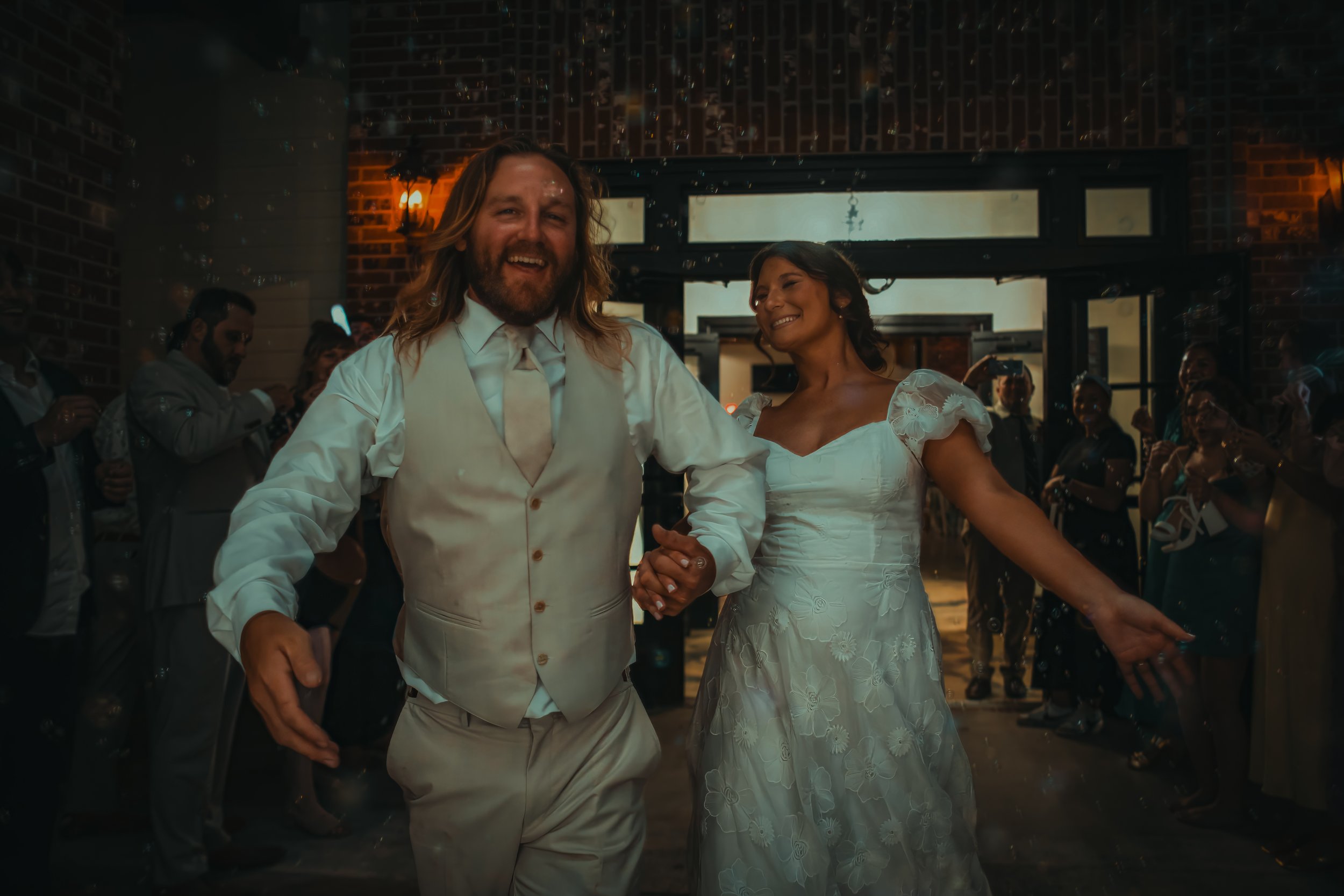 A couple dancing at a wedding reception, with guests in the background, in an indoor venue with brick walls and warm lighting.
