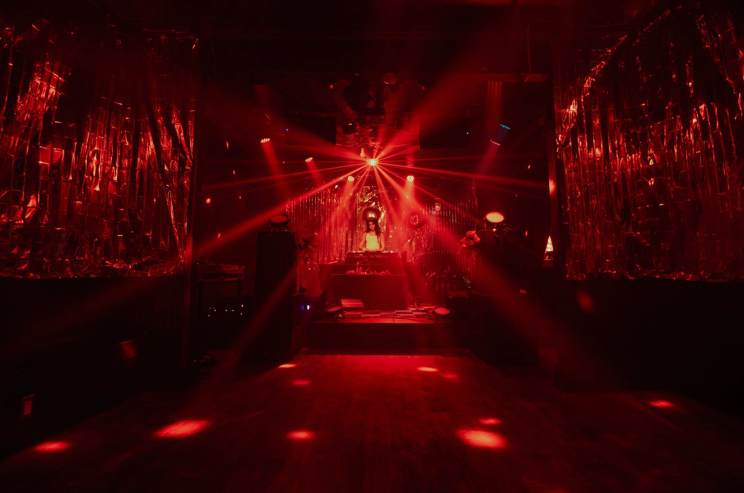 A nightclub stage illuminated with red lights, featuring a DJ booth at the center, surrounded by reflective metallic curtains and decorative flowers, with laser light beams projecting from the ceiling. L'objectif Photography: New Orleans Wedding, Eve