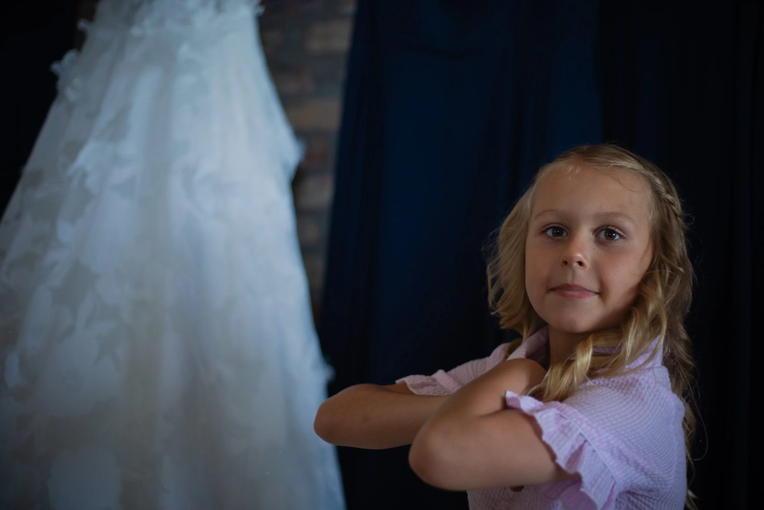 A young girl with blonde curly hair standing with her arms crossed, looking directly at the camera, wearing a pink dress, with part of a wedding dress on display next to her. L'objectif Photography: New Orleans Wedding, Event, and Portrait Photograph