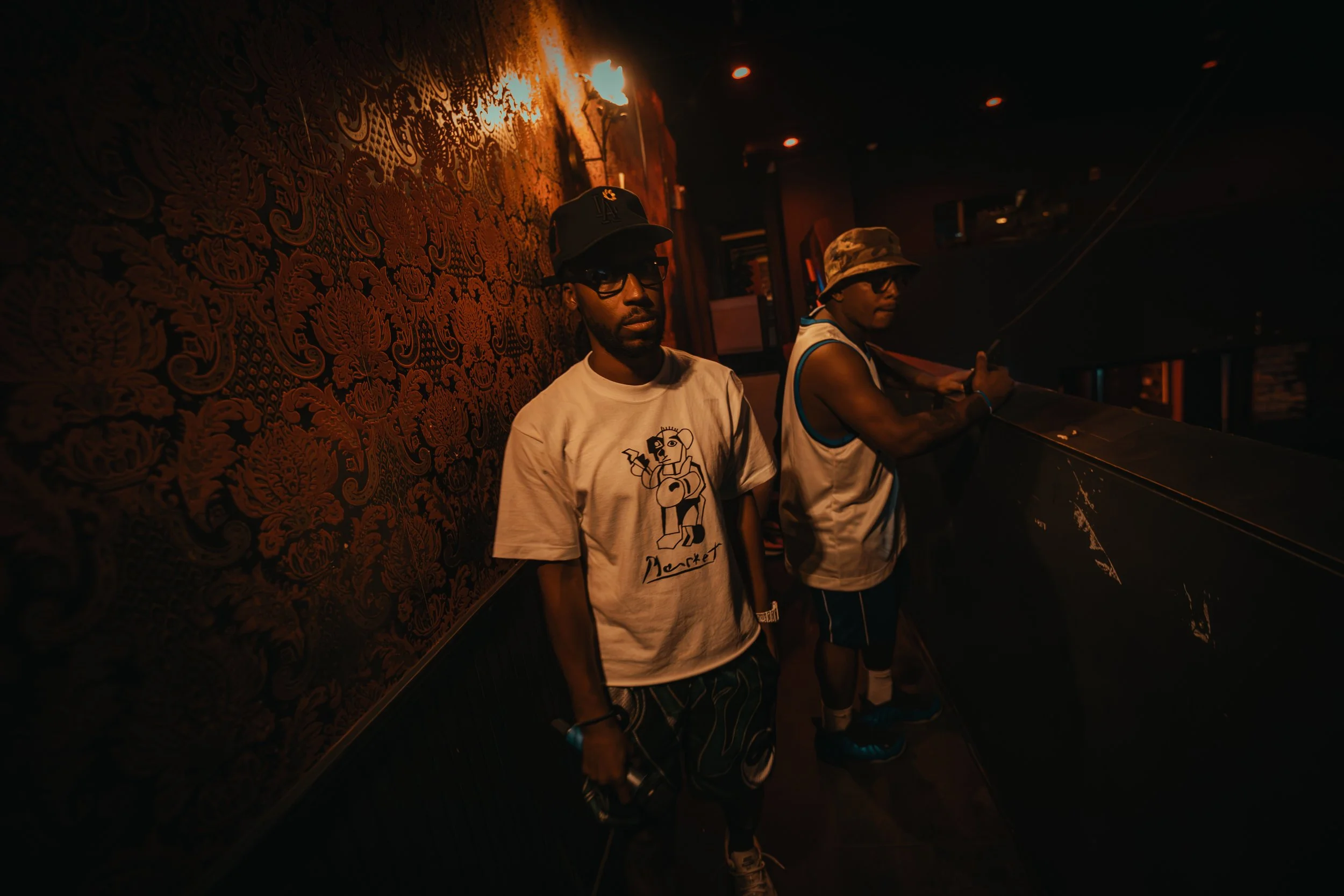 Two men standing in a dimly lit room next to a ornate wall with patterns, one wearing a baseball cap and glasses, the other wearing a camouflage bucket hat, both dressed casually.