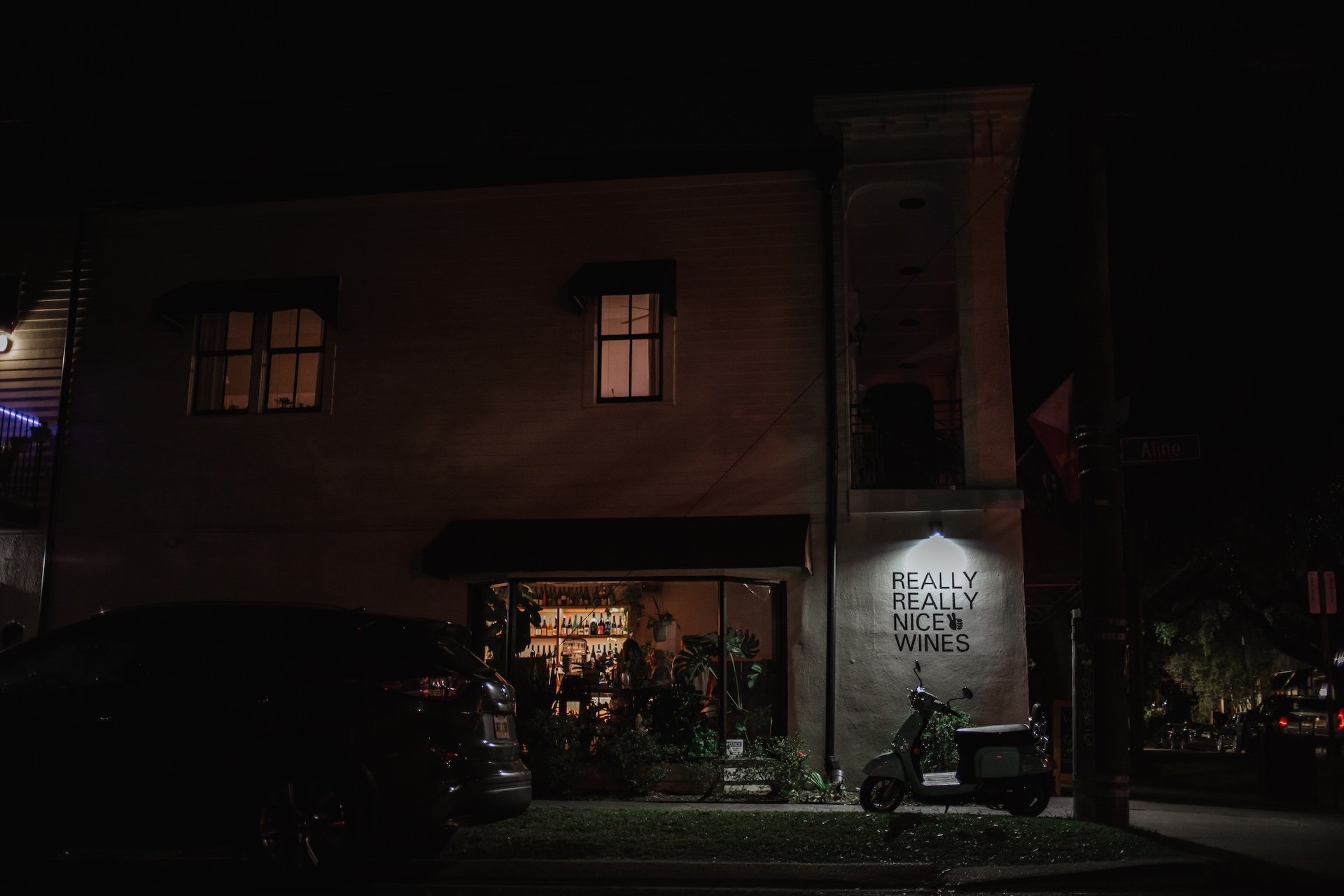 Night view of a two-story building with a bar or restaurant on the ground floor, illuminated from within. The sign on the building reads "Really Really Nice Wines." Cars and scooters are parked outside, and there is a streetlight near the sidewalk. L
