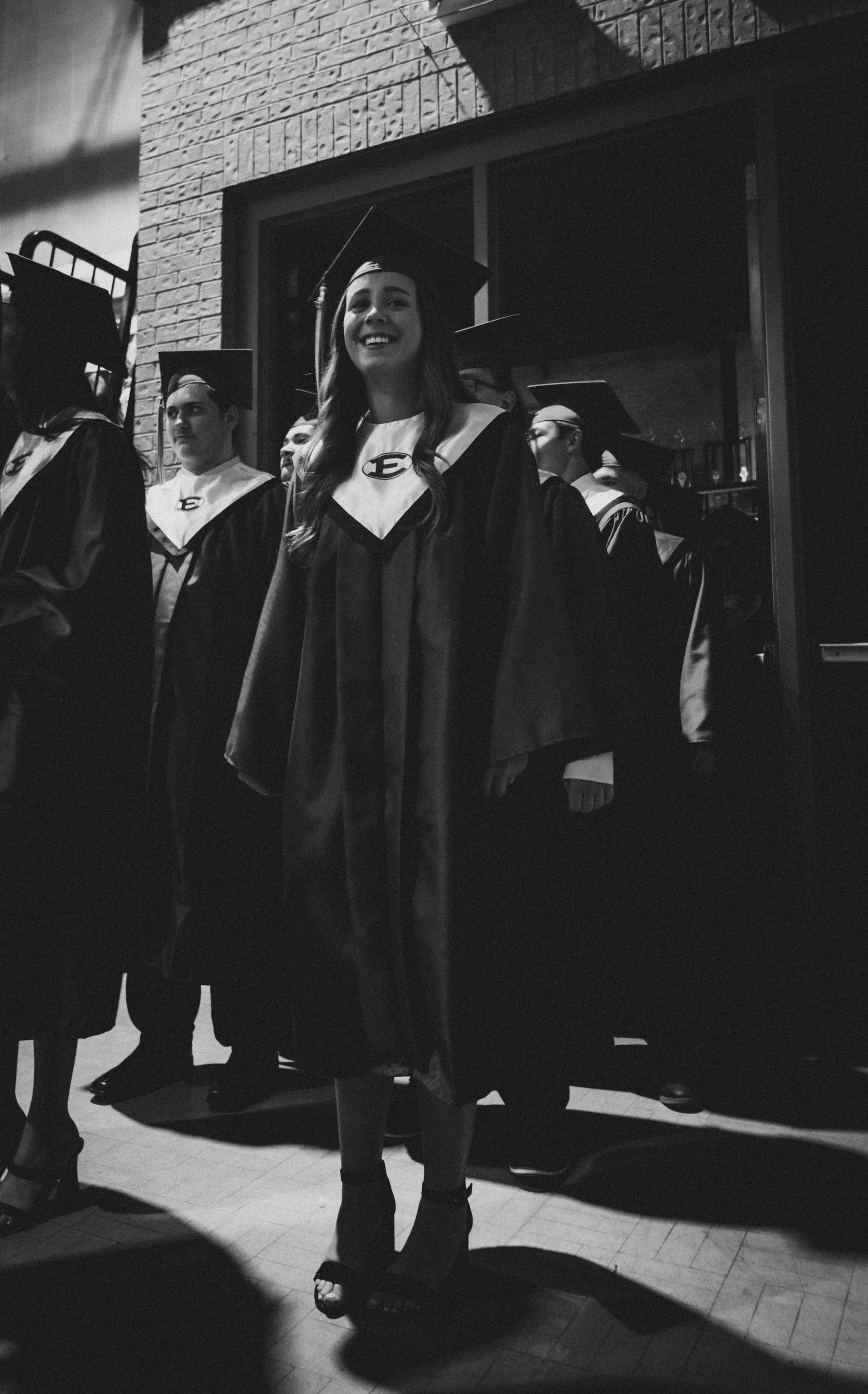 A young woman in a graduation cap and gown smiling, standing indoors with brick walls and other graduates around her.