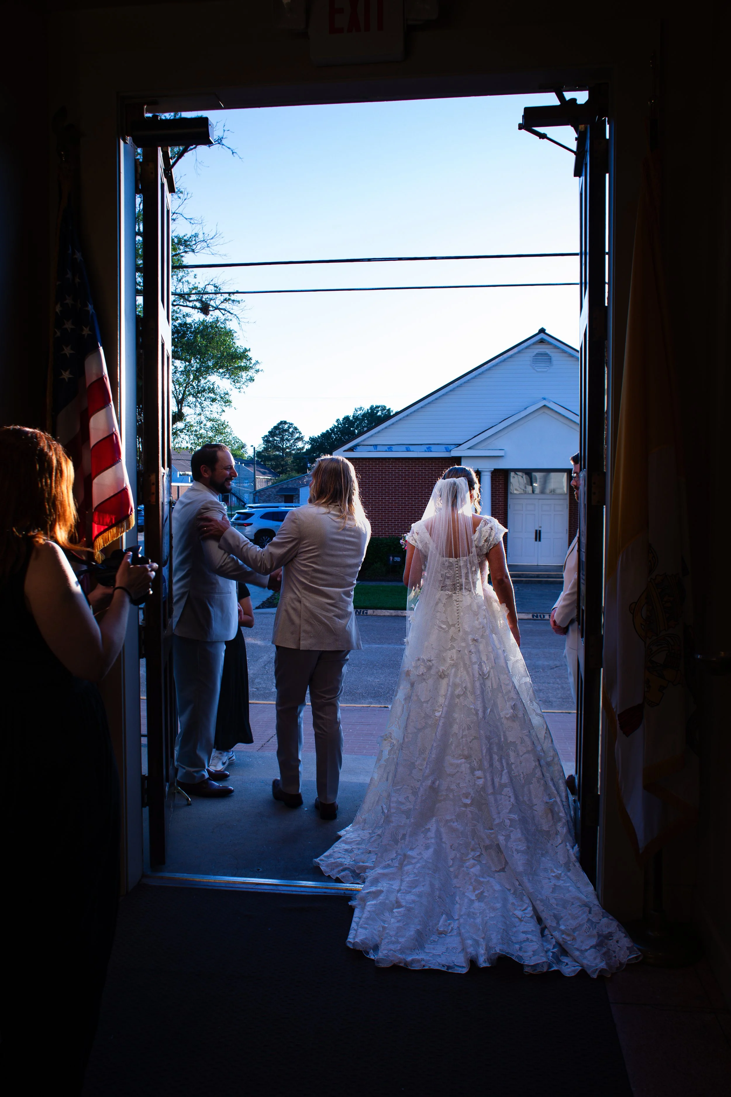 View from inside a building doorway showing a bride in a white gown and veil walking outside, with guests standing on either side, under a clear blue sky, during a wedding celebration.
