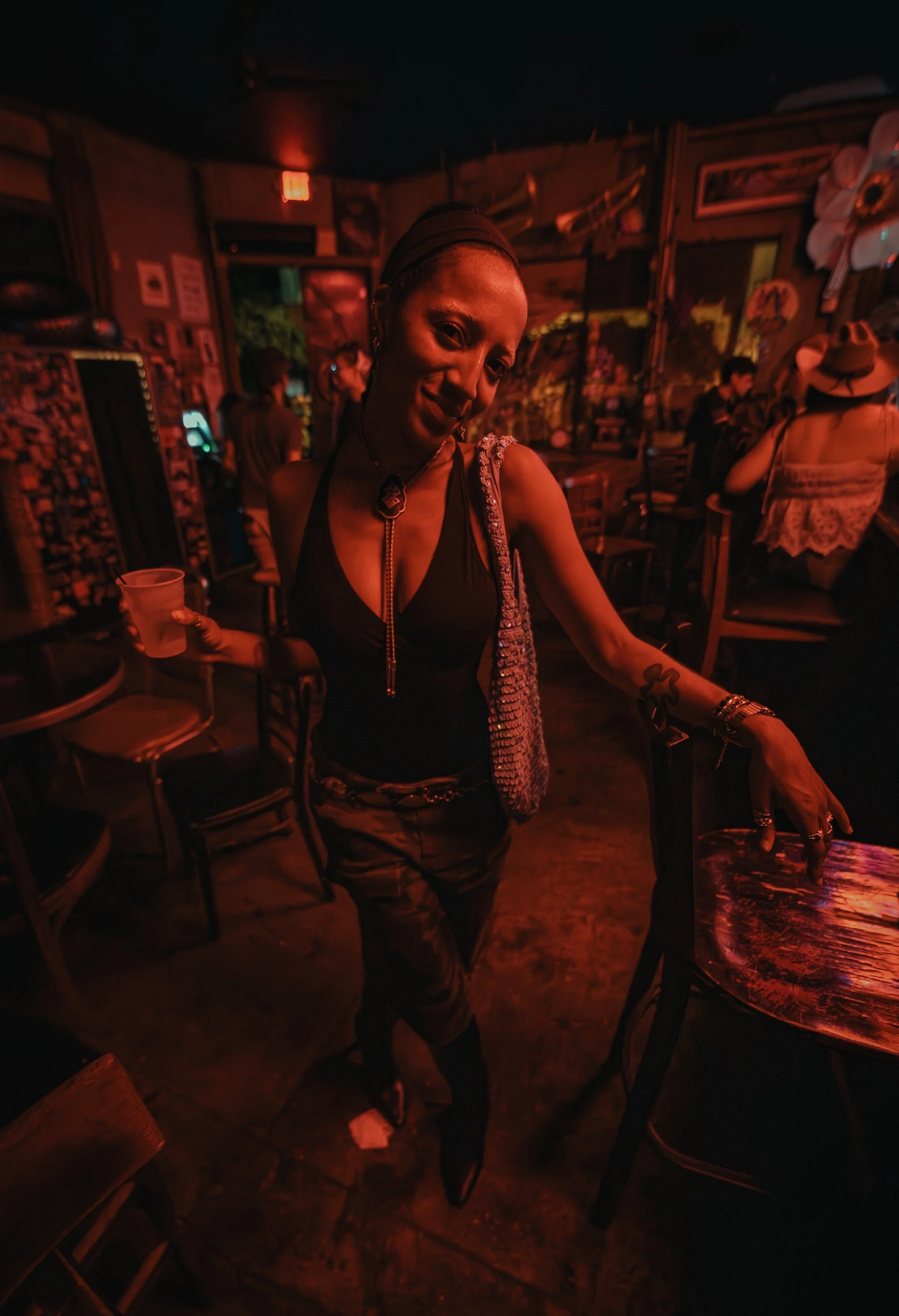 A woman in a dark, dimly lit bar or lounge, smiling and holding a drink. She is wearing a black top, camouflage pants, and jewelry. The background includes other people, chairs, and various wall decorations, with warm red lighting.