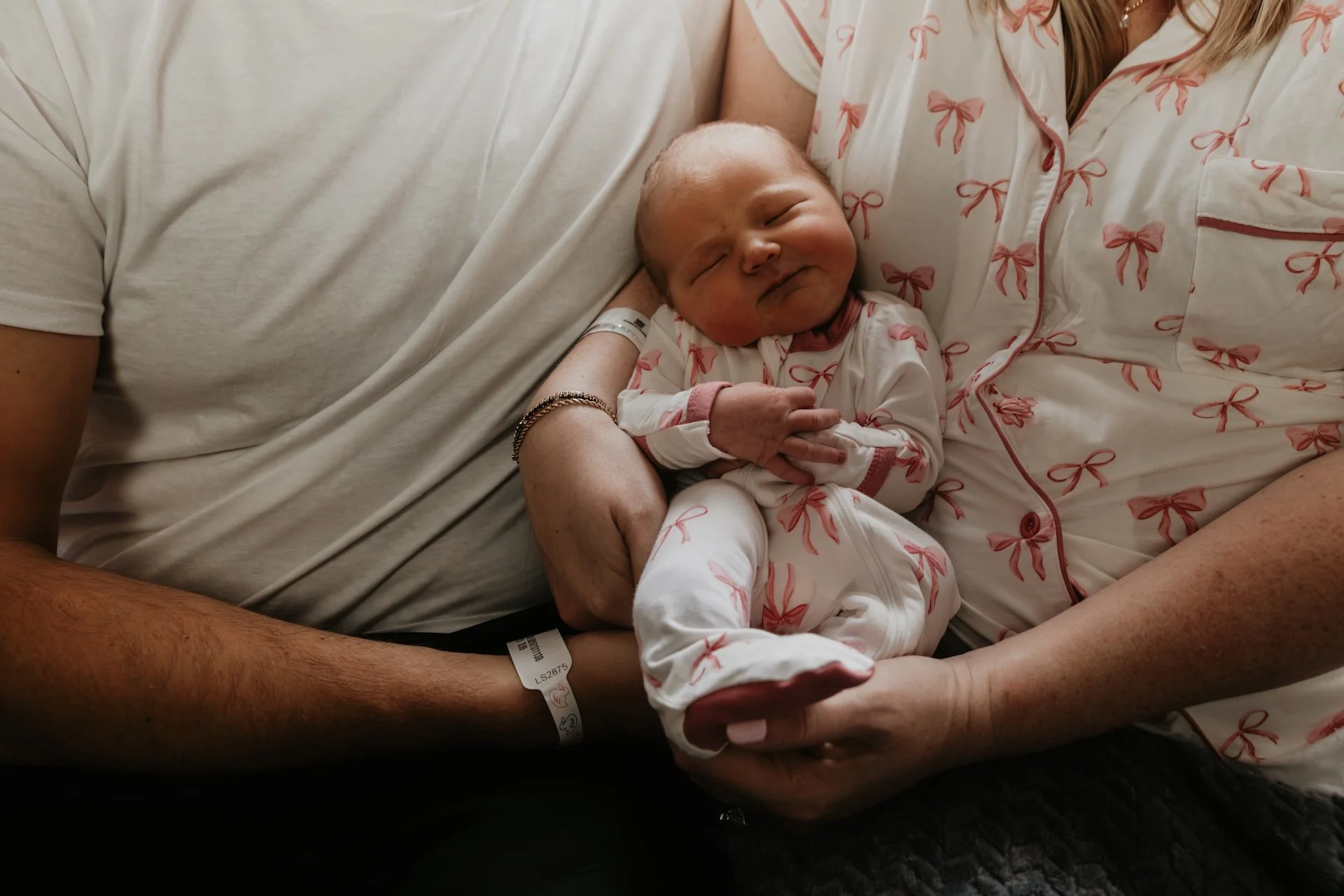 A newborn baby peacefully sleeping in the arms of a woman, likely the mother. The baby is dressed in white pajamas with pink bows, and the woman is wearing matching pajamas with the same bow pattern. The woman is sitting with the baby resting on her 