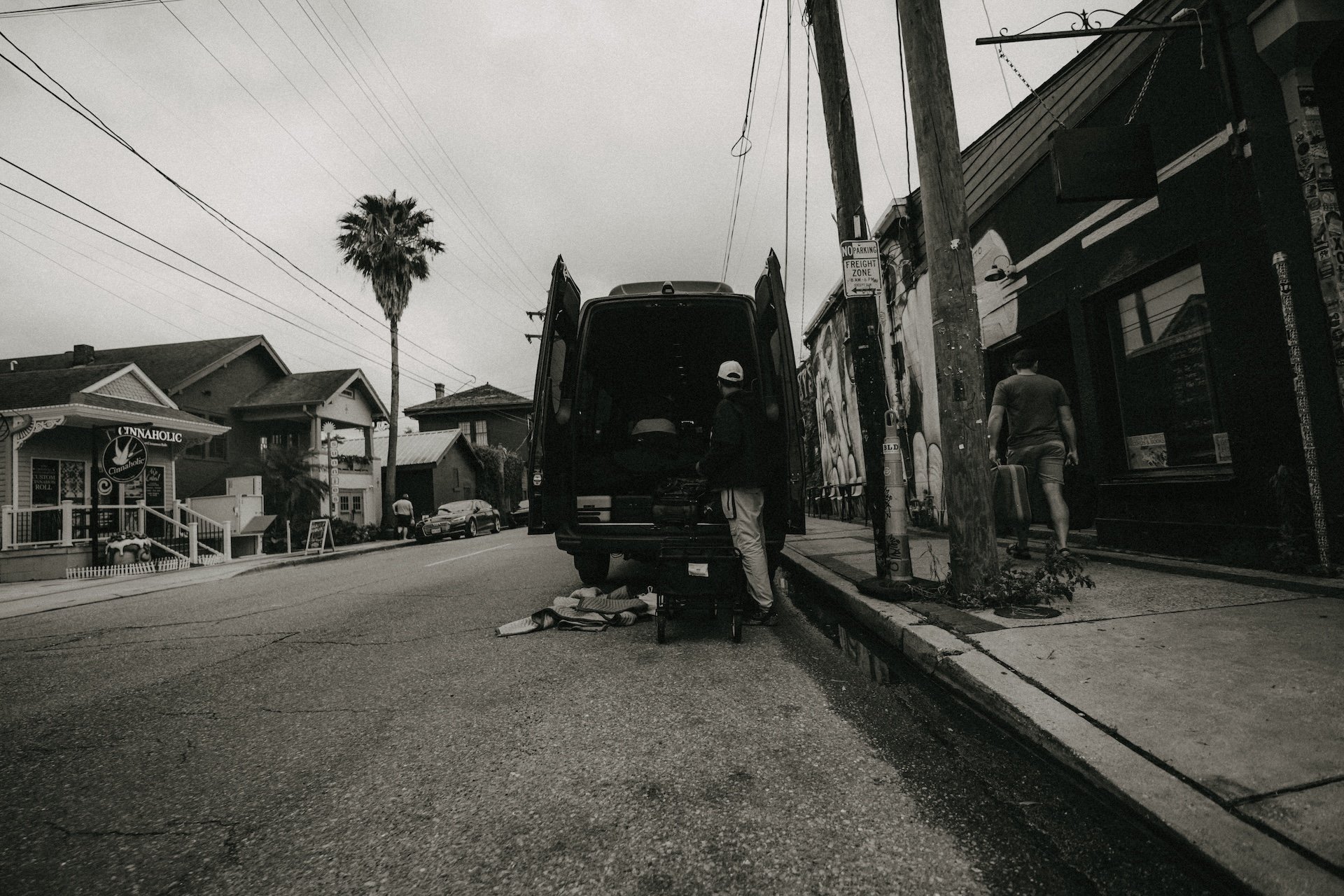 Man in a hat working on a van parked on the street, with an open back door and equipment nearby. A person with a briefcase is walking on the sidewalk. Residential houses, a palm tree, utility poles, and overhead wires are visible in the background. L