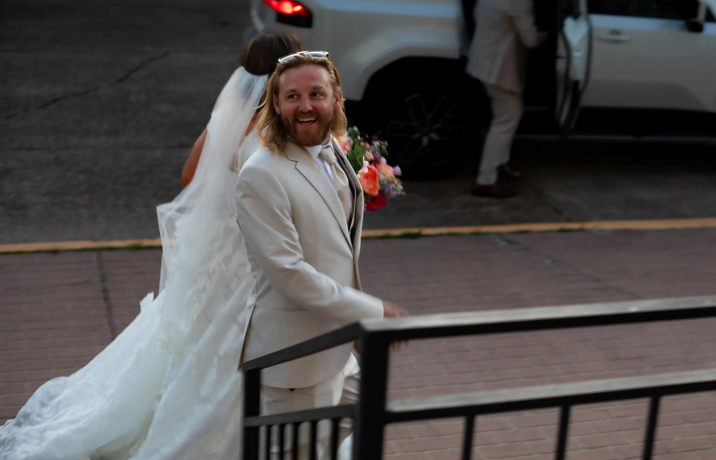 A man with long blond hair and a beard smiling while walking in a wedding suit holding a bouquet of flowers, with a woman in a wedding dress behind him, in front of a white vehicle. L'objectif Photography: New Orleans Wedding, Event, and Portrait Pho