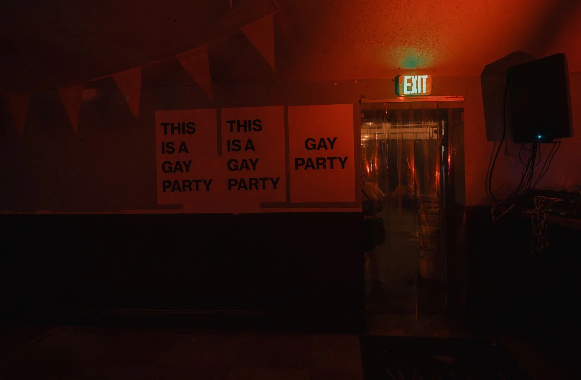 Dimly lit indoor space with red lighting, posters on the wall reading "THIS IS A GAY PARTY," a glass door, an exit sign, and a speaker on the wall. L'objectif Photography: New Orleans Wedding, Event, and Portrait Photographer