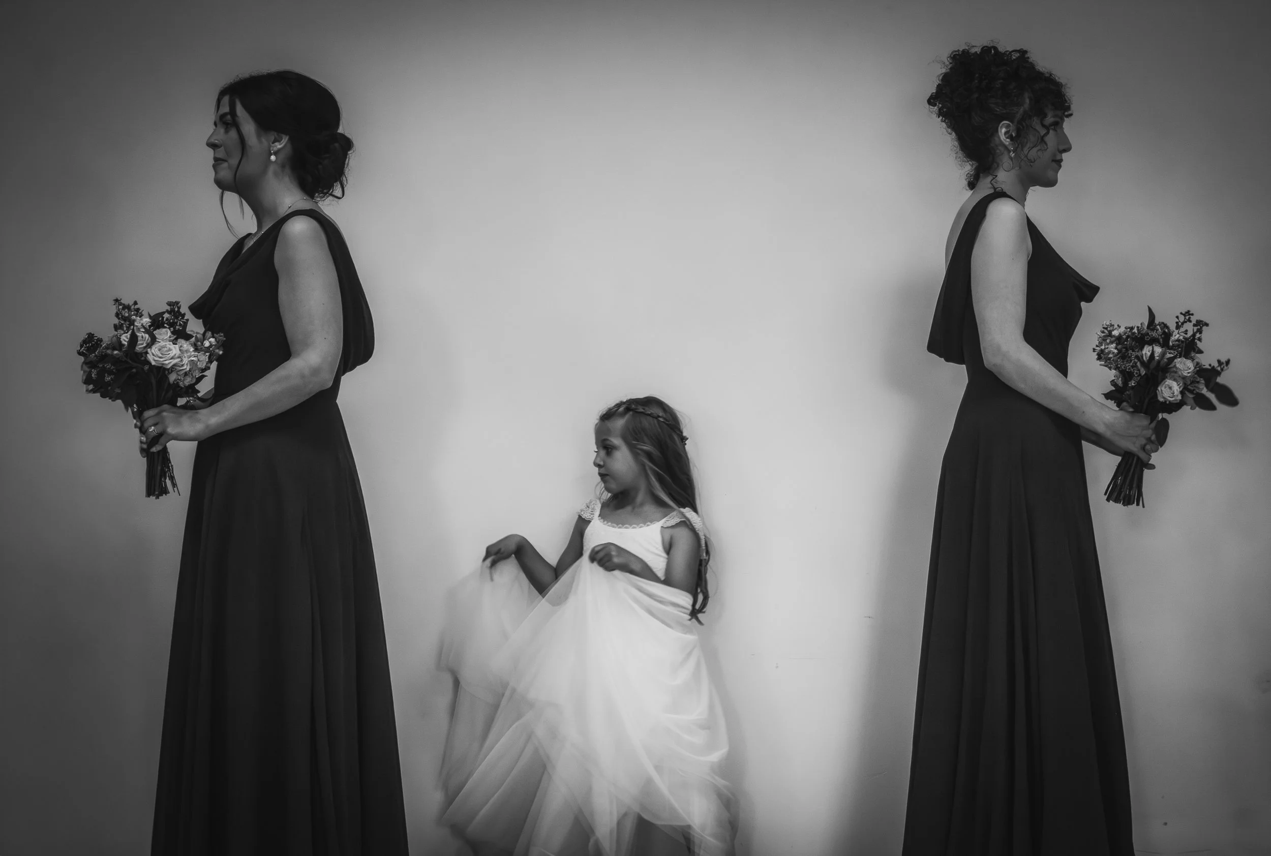 Two women in black dresses holding bouquets of flowers, standing opposite each other, with a young girl in a white dress between them, in front of a plain wall.