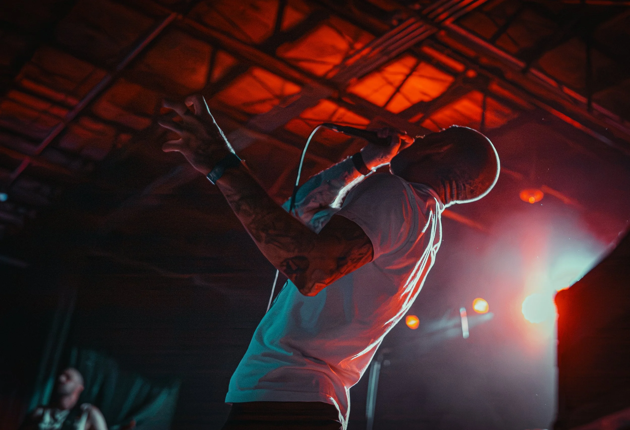 A musician on stage, wearing a cap and holding a microphone, performing under red-orange stage lighting with a dark background.