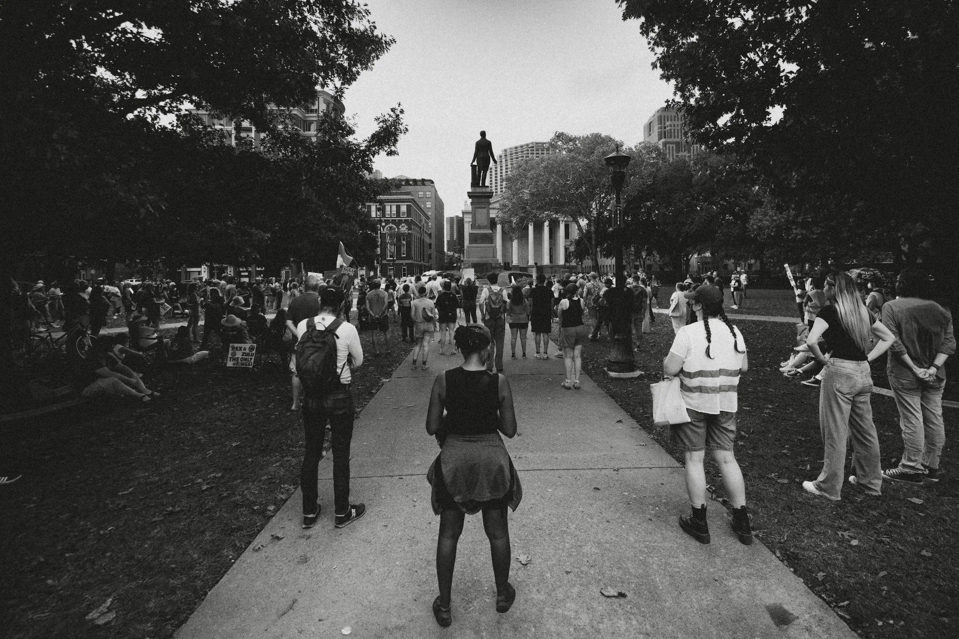 A black-and-white photo of a crowd of people gathered in a city park, with some sitting and others standing. A statue on a pedestal is in the background. Tall buildings surround the park, and trees frame the scene. L'objectif Photography: New Orleans