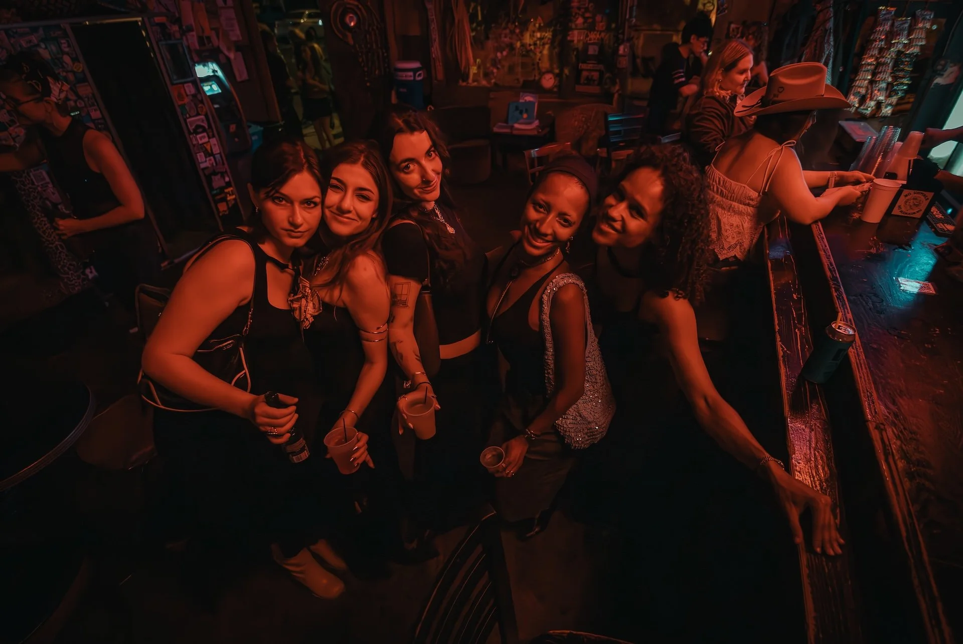 Group of five women standing together at a bar, smiling and holding drinks in a dimly lit, neon-colored setting. L'objectif Photography: New Orleans Wedding, Event, and Portrait Photographer