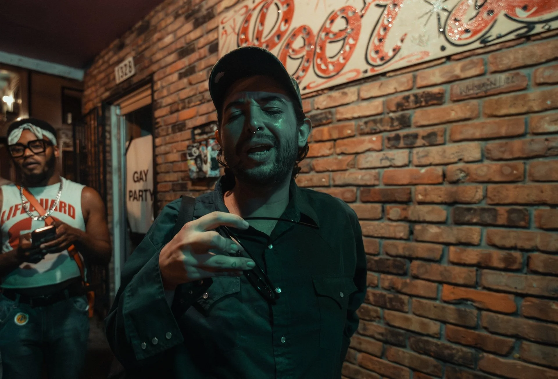 A woman with long dark hair, wearing a black cap and black jacket, is in front of an exposed brick wall at night, holding a pair of glasses. Behind her, a man with glasses, a bandana, and a tank top is looking at his phone. There is a sign on the bri
