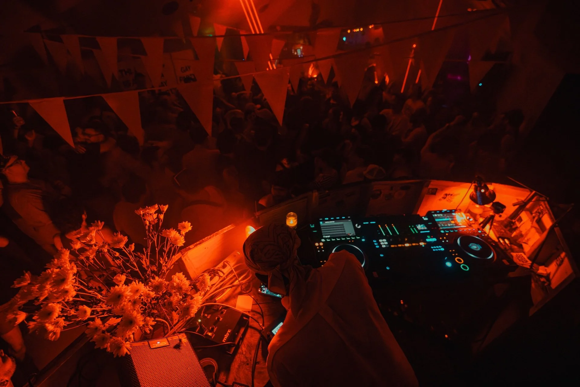 A DJ performing at a dark, red-lit nightclub with a crowd of people dancing and socializing. Flowers are placed on a table near the DJ setup. L'objectif Photography: New Orleans Wedding, Event, and Portrait Photographer