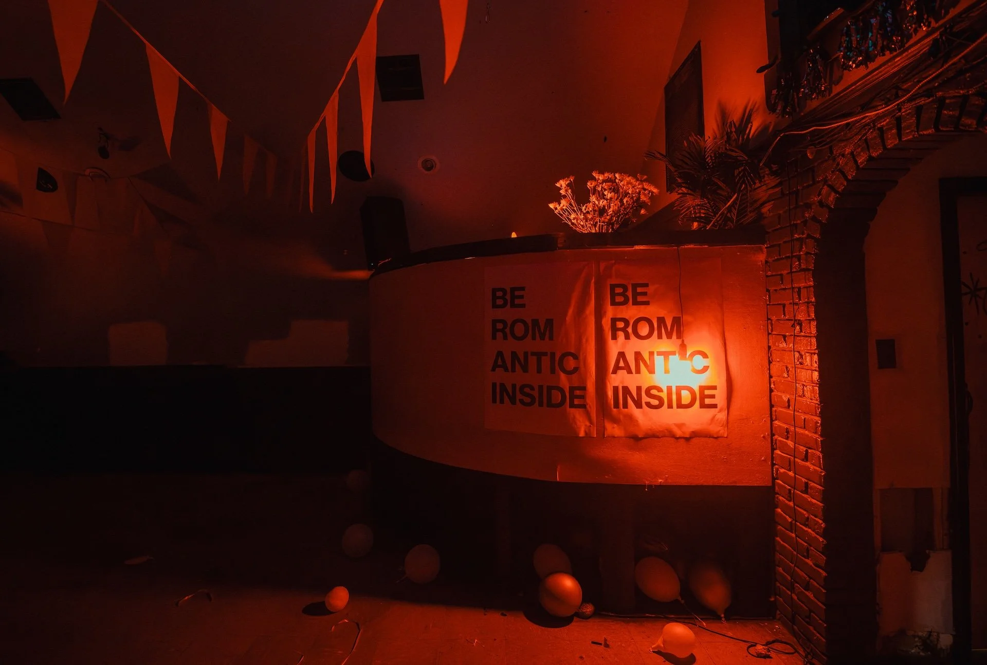 A dimly lit room with orange lighting, decorated for a celebratory event. There are orange triangular banners hanging from the ceiling, a curved wall with a sign that reads 'BE ROM ANTIC INSIDE' in black letters on orange paper, balloons scattered on