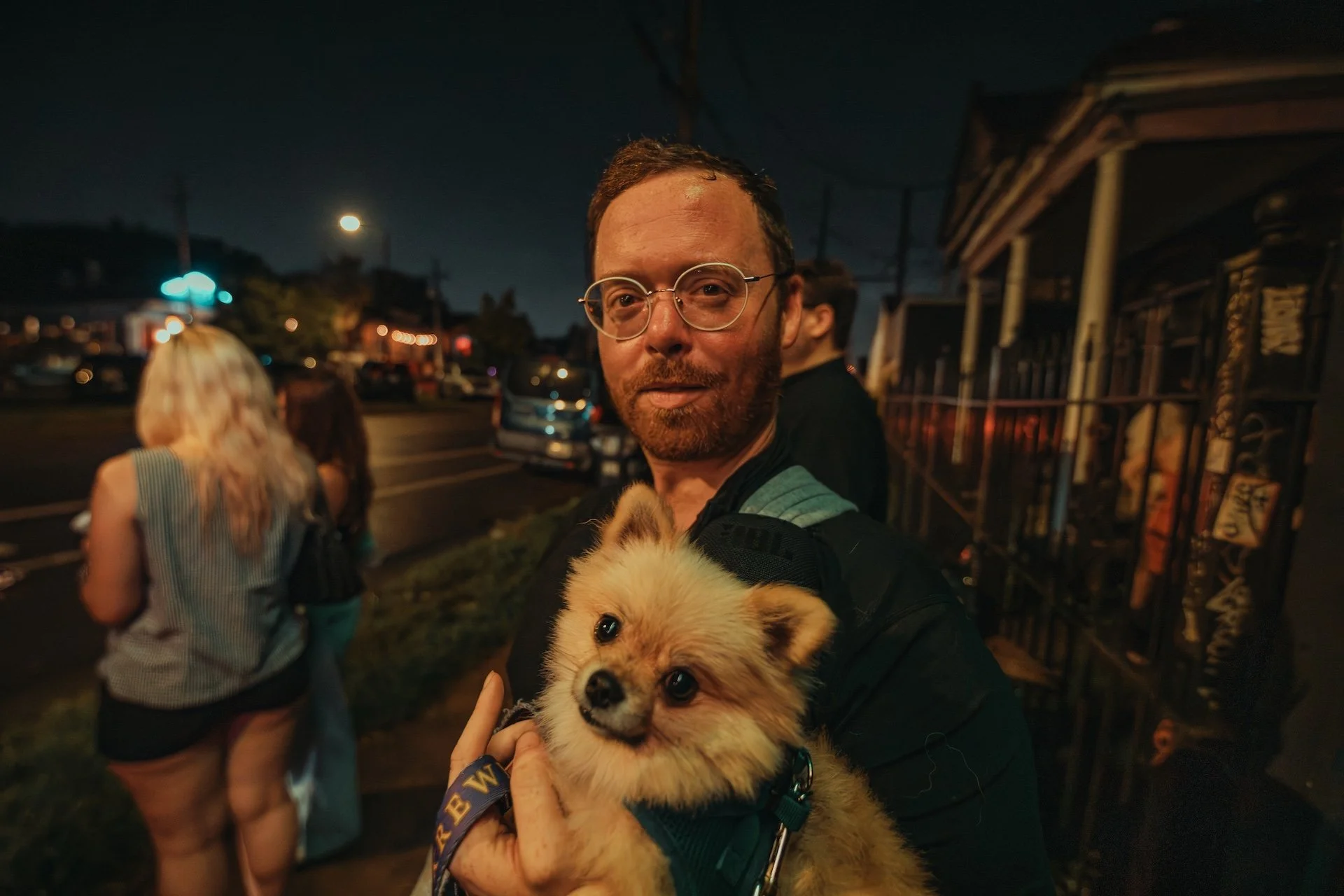 A man with glasses holding a fluffy tan and white puppy outdoors at night, with a woman with blonde hair and a striped top in the background. The scene is illuminated by streetlights and there are parked cars and a building nearby. L'objectif Photogr