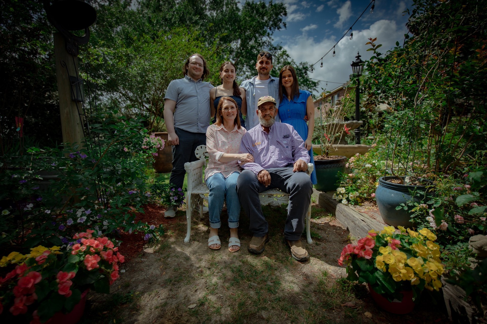 A multi-generational family is gathered in a lush garden filled with colorful flowers. There are seven people, including an older man and woman seated on a white garden bench, surrounded by potted plants and greenery under a partly cloudy sky, with s