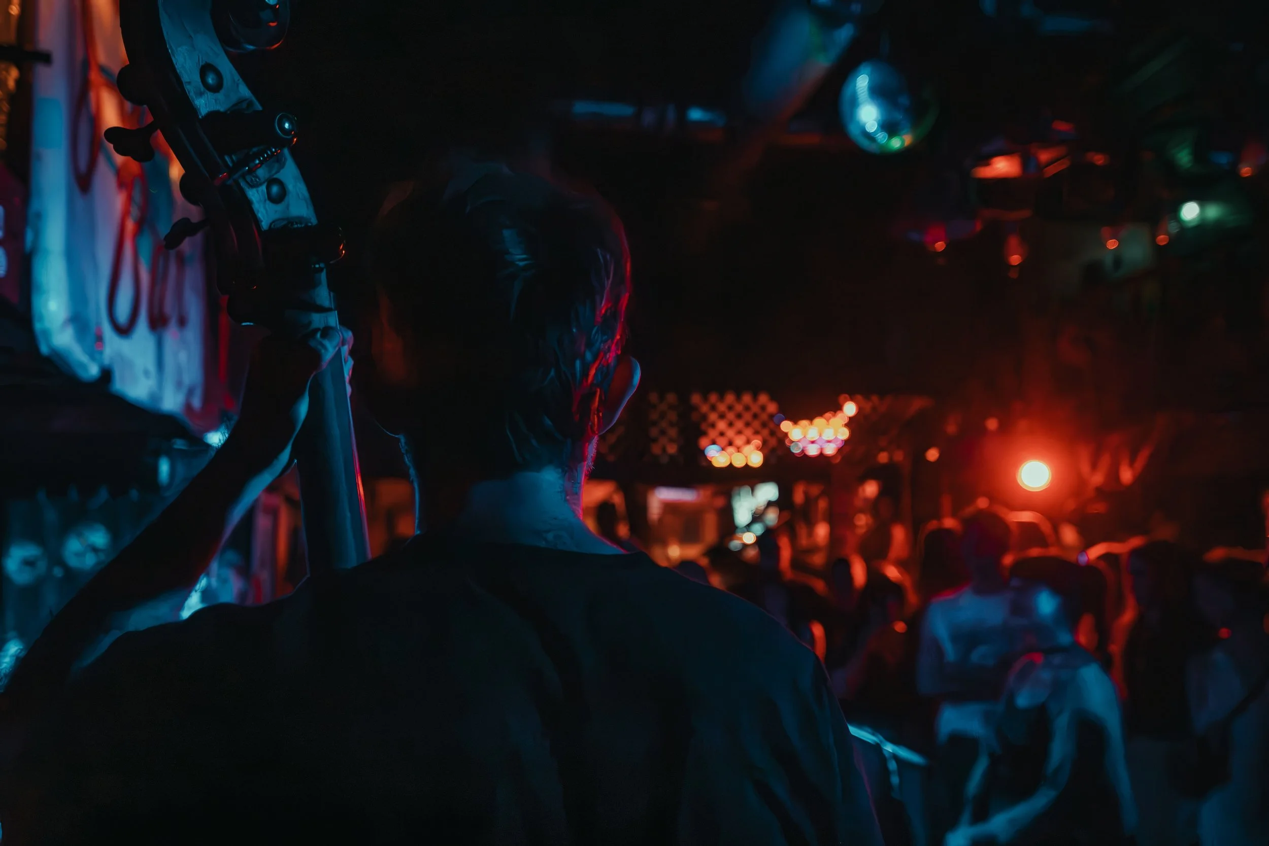 A dark scene with neon and red lighting, showing a person wearing a baseball cap and jacket, with their back to the camera among a crowd of people and cars. L'objectif Photography: New Orleans Wedding, Event, and Portrait Photographer