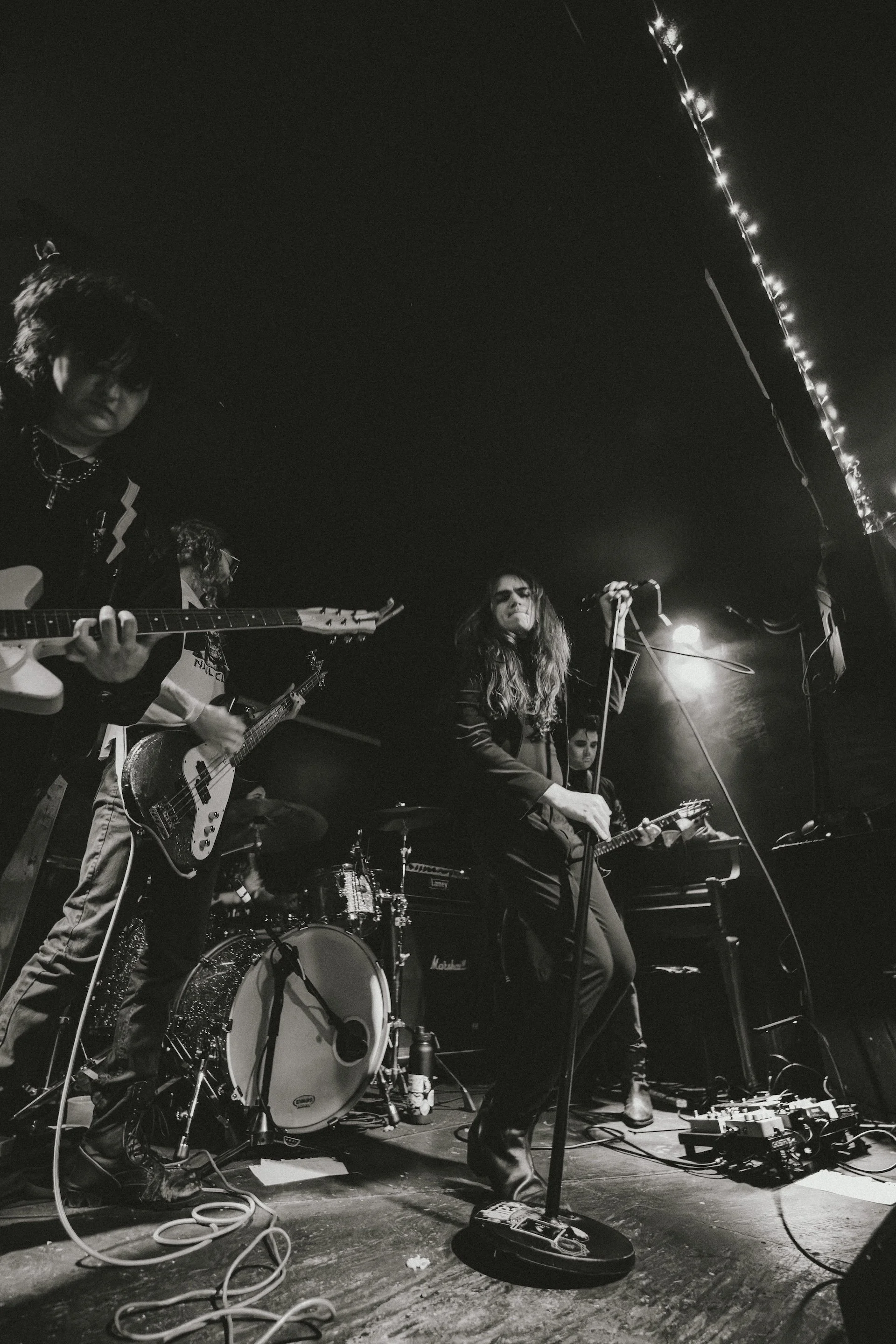 A black-and-white photo of a live musical performance. Four musicians are on stage, with one male guitarist on the left, a female singer in the middle, and other band members visible in the background. The stage is dimly lit with string lights and various musical equipment and cables on the floor.