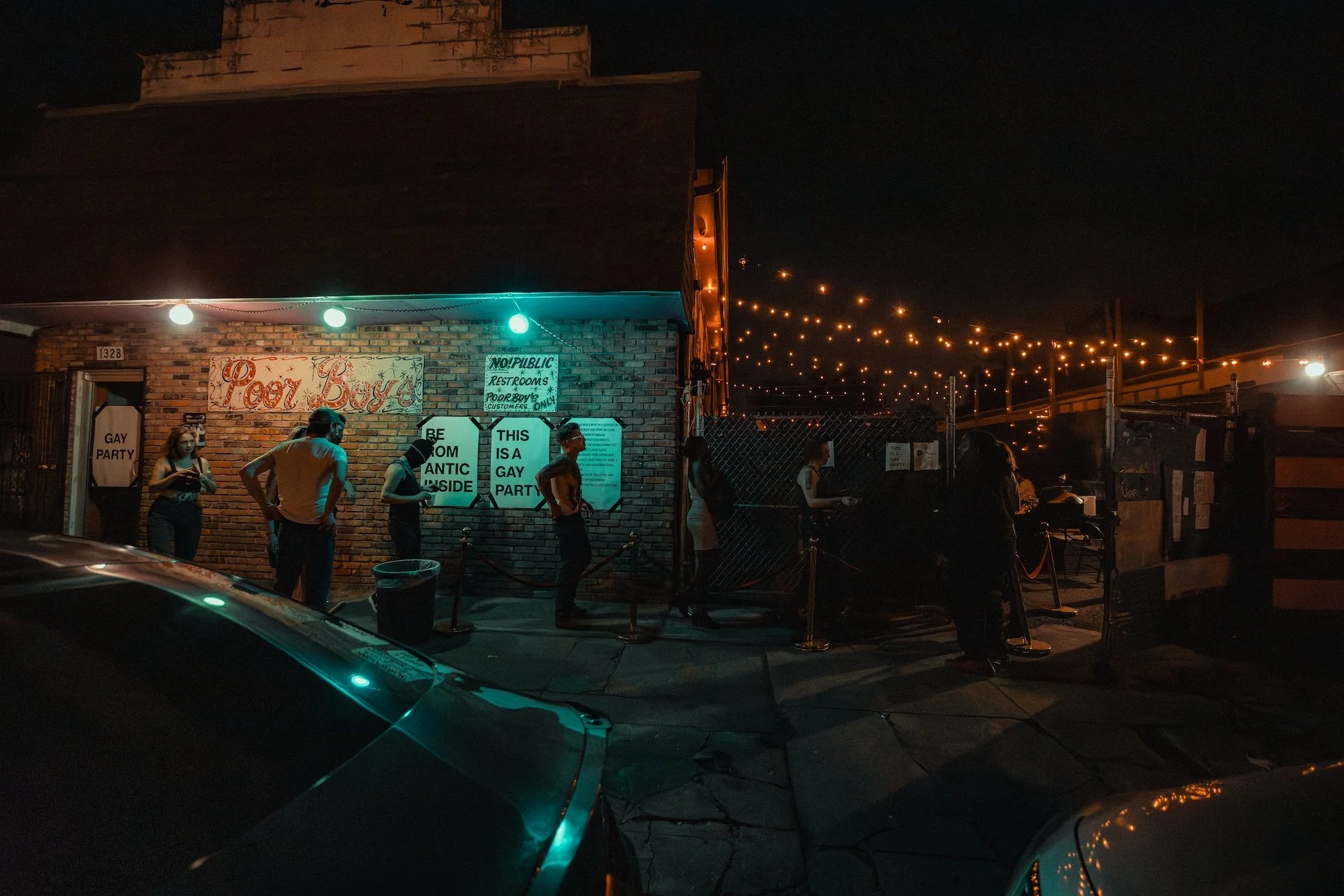 Nighttime scene outside a brick building with a sign that reads 'Food Box.' Several people are standing in line or waiting, with some signs indicating it is a gay-friendly event. String lights hang above, illuminating the area gently, and there are c